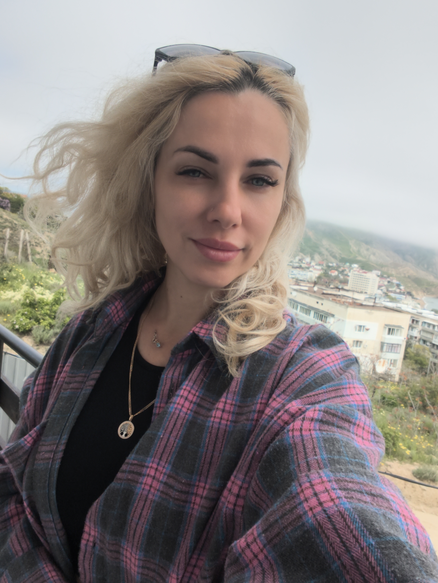 A fair-skinned woman with blonde, shoulder-length hair is centered in the frame, posing for a selfie-style photo. She has sunglasses perched on her head and is wearing a black top and a grey and pink plaid flannel shirt. A gold necklace with a circular pendant is visible around her neck.
The woman is on a balcony, with a blurred landscape visible in the background. In the distance, a town or city is partially visible, with buildings and a hazy, overcast sky above. Vegetation is visible on the hills or slopes in the background. The photo is taken in the city of Орджоникидзевский поселковый совет, Ukraine.