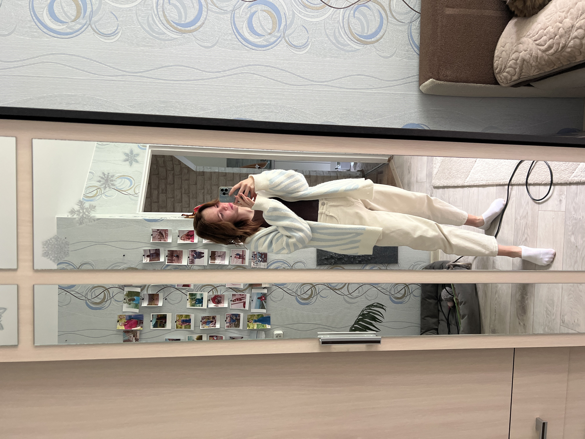 The scene depicts a young woman taking a mirror selfie in what appears to be a bedroom or dressing area within a domestic residence, located in the city of Chaikovsky, Russia.
The central subject is a light-skinned woman, facing left and holding a light-colored smartphone with her right hand, seemingly taking a photo of her reflection. She has shoulder-length brown hair, a small red festive hair accessory, and is looking intently at the phone's screen. She is wearing a dark brown long-sleeved top, a loose-fitting white cardigan with light blue vertical stripes, and light-colored (possibly off-white or cream) relaxed-fit pants. Her feet, visible below the pants, are clad in white ankle socks on a light wooden floor. A thin black cable is coiled on a light-colored shag rug positioned on the floor to her left.
The woman is standing in front of a tall wardrobe featuring three mirror panels framed by light-colored wood. The leftmost mirror panel has several small, rectangular photos (resembling instant camera prints) taped to its surface, depicting various individuals and groups in different settings. A two-pin white electrical outlet is reflected on the wall to the left of these panels. Further left, a light wooden wardrobe with a metal handle is visible. A dark grey pet bed with a toy (a pink ball with white string) is reflected on the floor, occupied by a partially visible dark-colored pet, possibly a dog or cat. The reflection also shows a glimpse of a large green plant leaf.
To the right of the woman, beyond the mirrored wardrobe, the room's wall is adorned with light grey wallpaper featuring an abstract pattern of blue, gold, and grey concentric circles and wavy lines. Several white snowflake-shaped decorations are affixed to this wall near the ceiling line. A bed with a brown upholstered headboard and a decorative patterned pillow is visible on the far right, with a ginger or tabby cat resting on it. Further in the background, reflected in the mirrors on the far left, a doorway leads to an adjacent room with a visible light-colored brick-patterned wall.