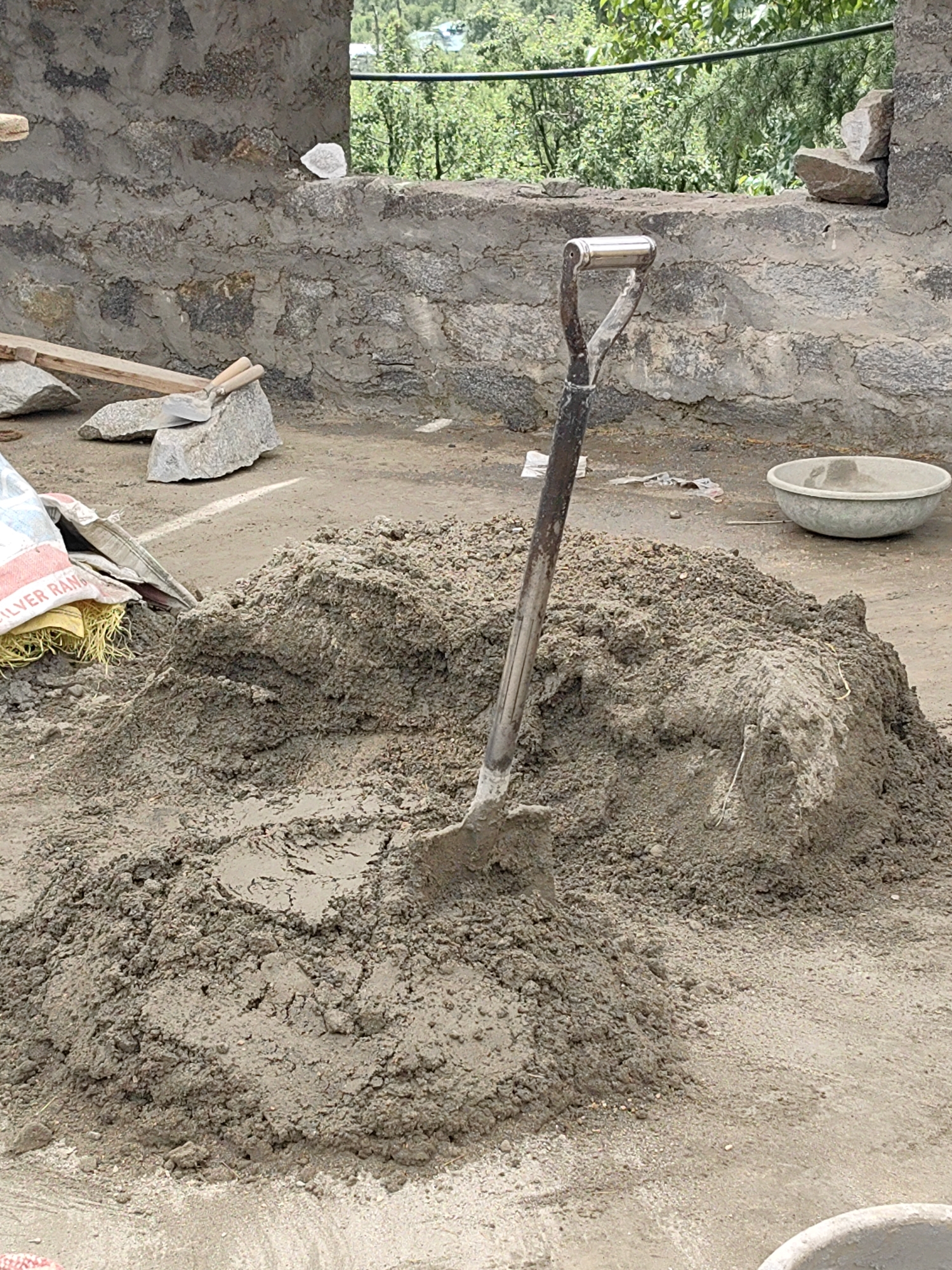 In Vashisht, India, a construction site is depicted with a large pile of cement mixture in the foreground. A shovel is embedded in the pile, indicating recent or ongoing work. To the right of the pile, a shallow, round concrete basin is visible. In the background, a rough stone wall forms the boundary of the construction area, with greenery and trees beyond. The scene suggests a construction project in progress, likely for building or renovation purposes.