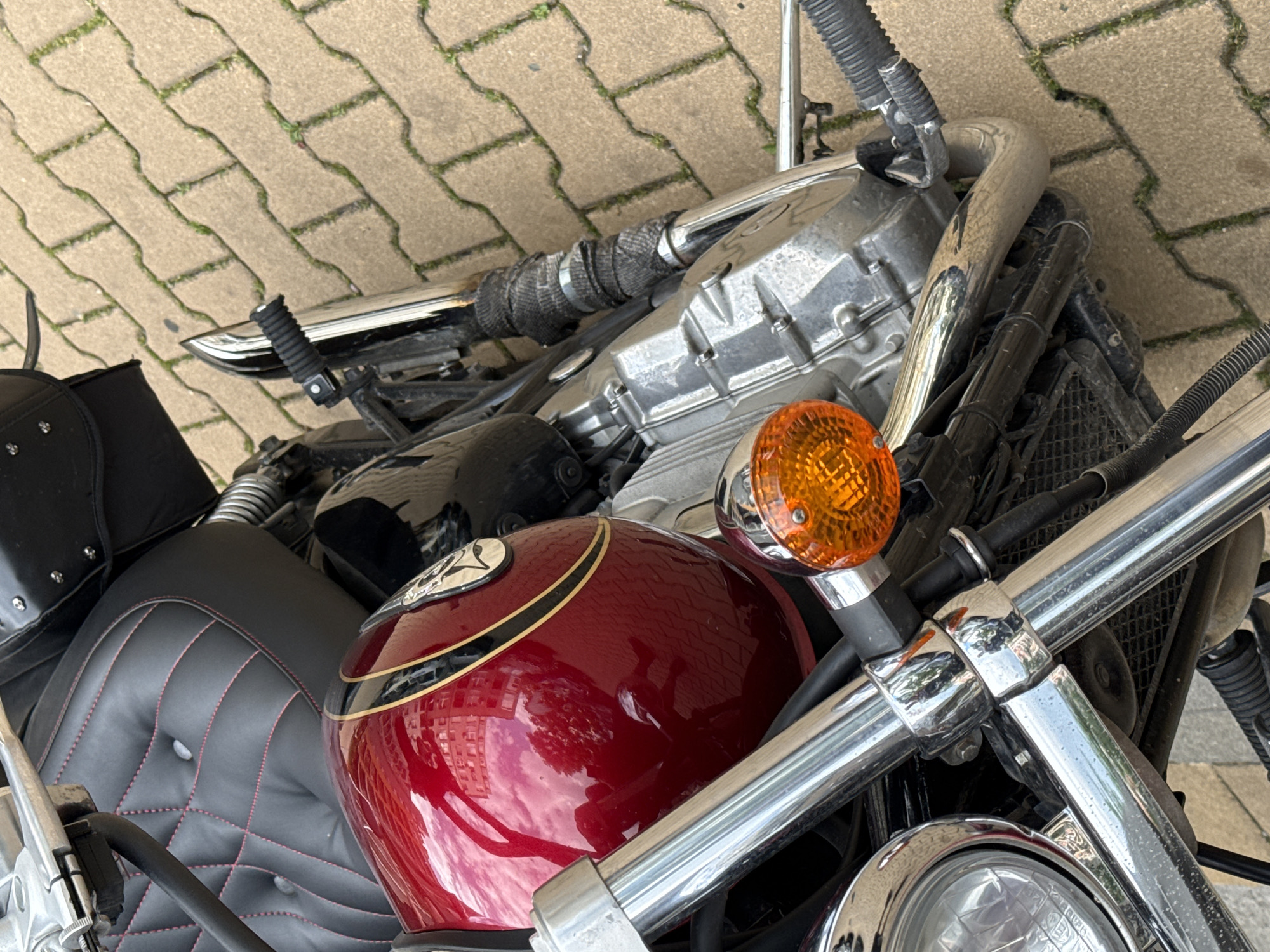 This image, captured in Sofia, Bulgaria, displays a stationary cruiser-style motorcycle viewed from an elevated, downward perspective. The motorcycle is predominantly black with extensive chrome detailing. Its fuel tank is a glossy, metallic maroon, adorned with gold and black pinstriping, and vividly reflects parts of a multi-story building with rectangular windows, a cloudy sky, and surrounding tree foliage. The black seat features a diamond-quilted pattern with contrasting red stitching and four visible silver button tufts. Prominent chrome elements include the handlebars, front forks, a round headlight housing, and a section of the exhaust pipe. The front right turn signal is a round, amber unit. The polished silver engine casing is visible on the right side, adjacent to a segment of the exhaust pipe that is wrapped in dark grey heat-resistant material. A black rubberized foot peg is also present on the right side. The motorcycle is positioned on a paved ground surface constructed from light-colored, rectangular interlocking concrete pavers, with green moss or weeds growing within the joints. No individuals are visible, and the scene is illuminated by natural daylight.