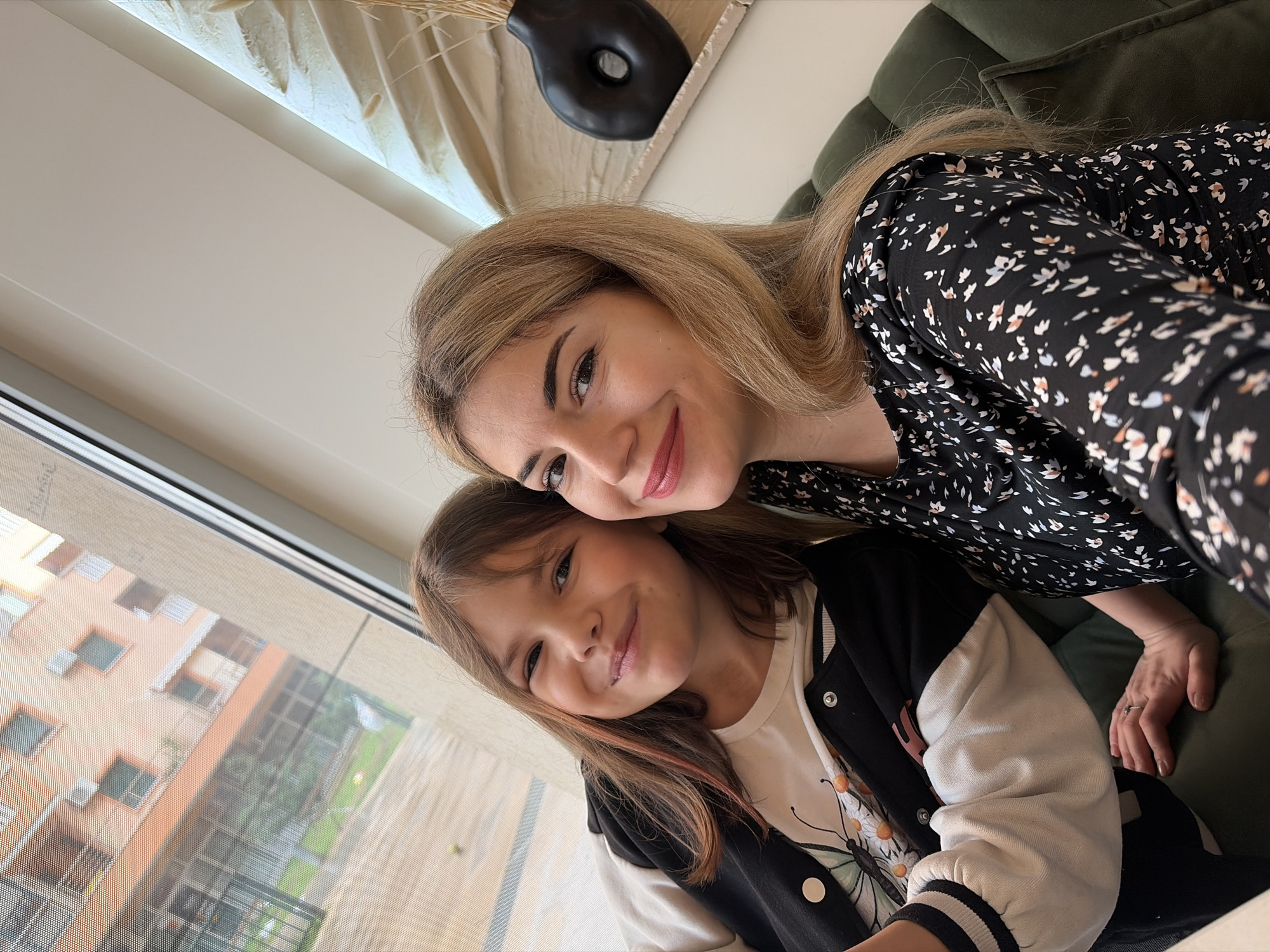 An indoor selfie depicts two females seated on a dark green upholstered couch or booth. The subject on the right, an adult female with long blonde hair, wears a black long-sleeved garment with a small, scattered floral pattern and dark pink lipstick. Her left hand, featuring a ring on the ring finger, rests on the green upholstery. Adjacent to her right, a beige textured wall hanging serves as a backdrop for a black, organic-shaped vase containing dried reeds. The subject on the left, a younger female with long brown hair including reddish highlights, smiles at the camera while wearing a white t-shirt with a colorful butterfly motif under a black varsity-style jacket with white sleeves. Both subjects are smiling directly at the camera. Behind them, a large window provides a view through a fine mesh screen to the exterior. Visible outdoors is a multi-story building with a peach-toned facade, featuring air conditioning units and balconies, alongside green foliage and a paved ground area. This scene is located in Budva, Montenegro.