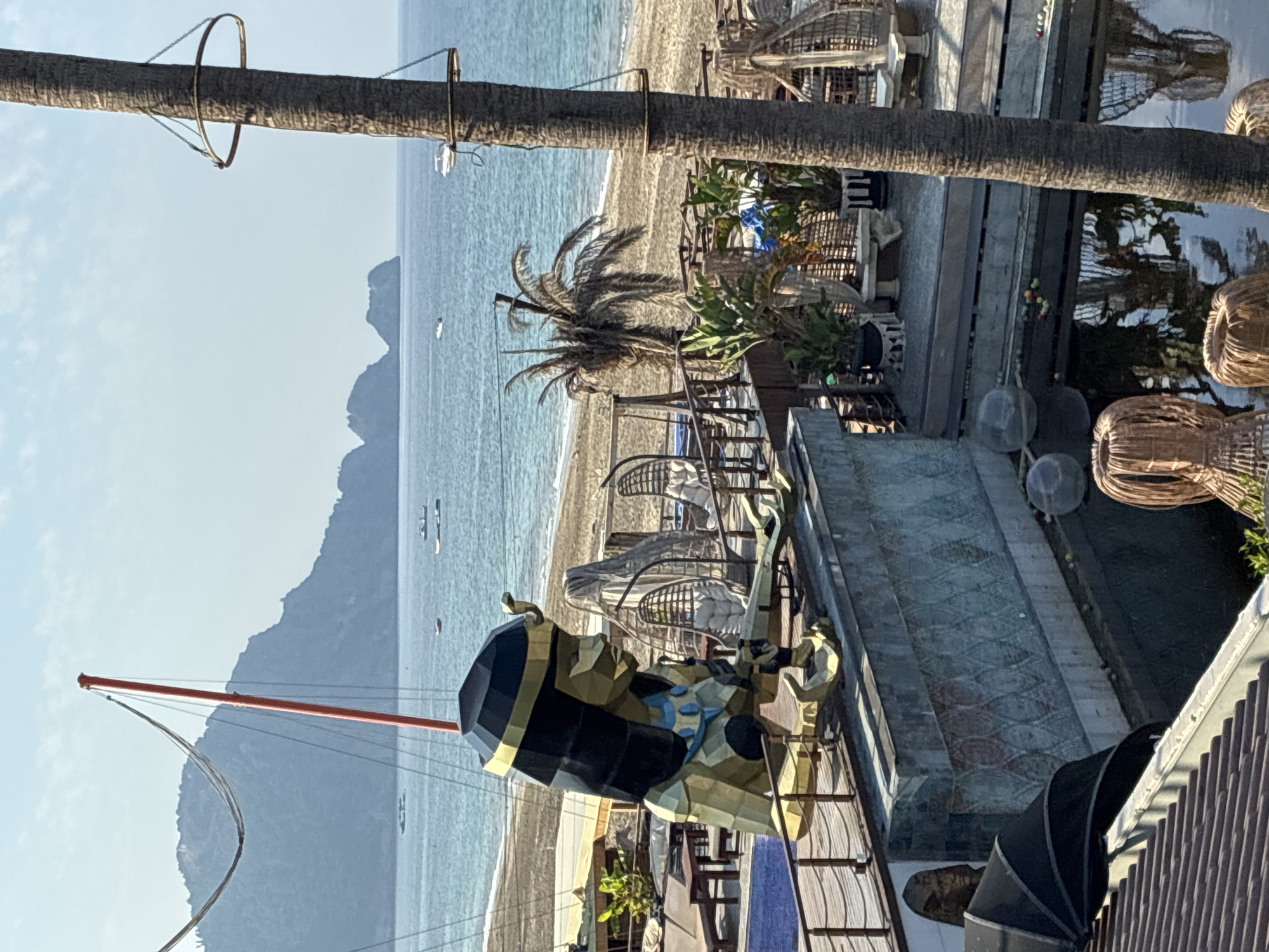 A large, geometric, golden statue of an Egyptian figure, likely Cleopatra, is positioned on a concrete ledge overlooking a beach and the Mediterranean Sea in Alanya, Turkey. The statue is gold and black, with a headdress and decorative collar. Behind the statue, beach chairs, lounge chairs, and wicker seating are visible, suggesting a resort or outdoor dining area. The beach is sandy, and the sea is a clear blue with several small boats in the distance. A tall palm tree trunk with metal rings attached is in the foreground on the right. Mountains are visible in the background. The water in the foreground appears to be a pond or pool with reflections of the surroundings.