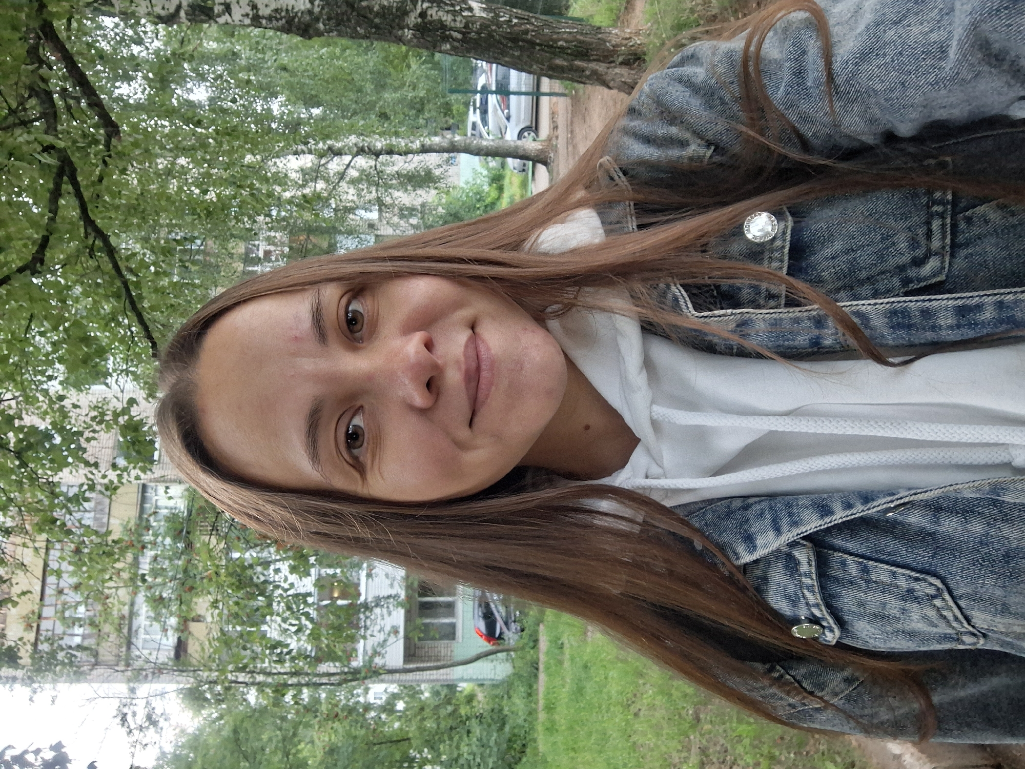 An outdoor medium shot shows a young woman with long brown hair, smiling at the camera. She is wearing a denim jacket over a white hoodie. She has light brown eyes and fair skin with some blemishes on her cheeks. In the background, there is a multi-story apartment building, trees, green grass, and parked cars, consistent with a residential area in Chaikovsky, Russia. The lighting suggests it is daytime, possibly overcast.