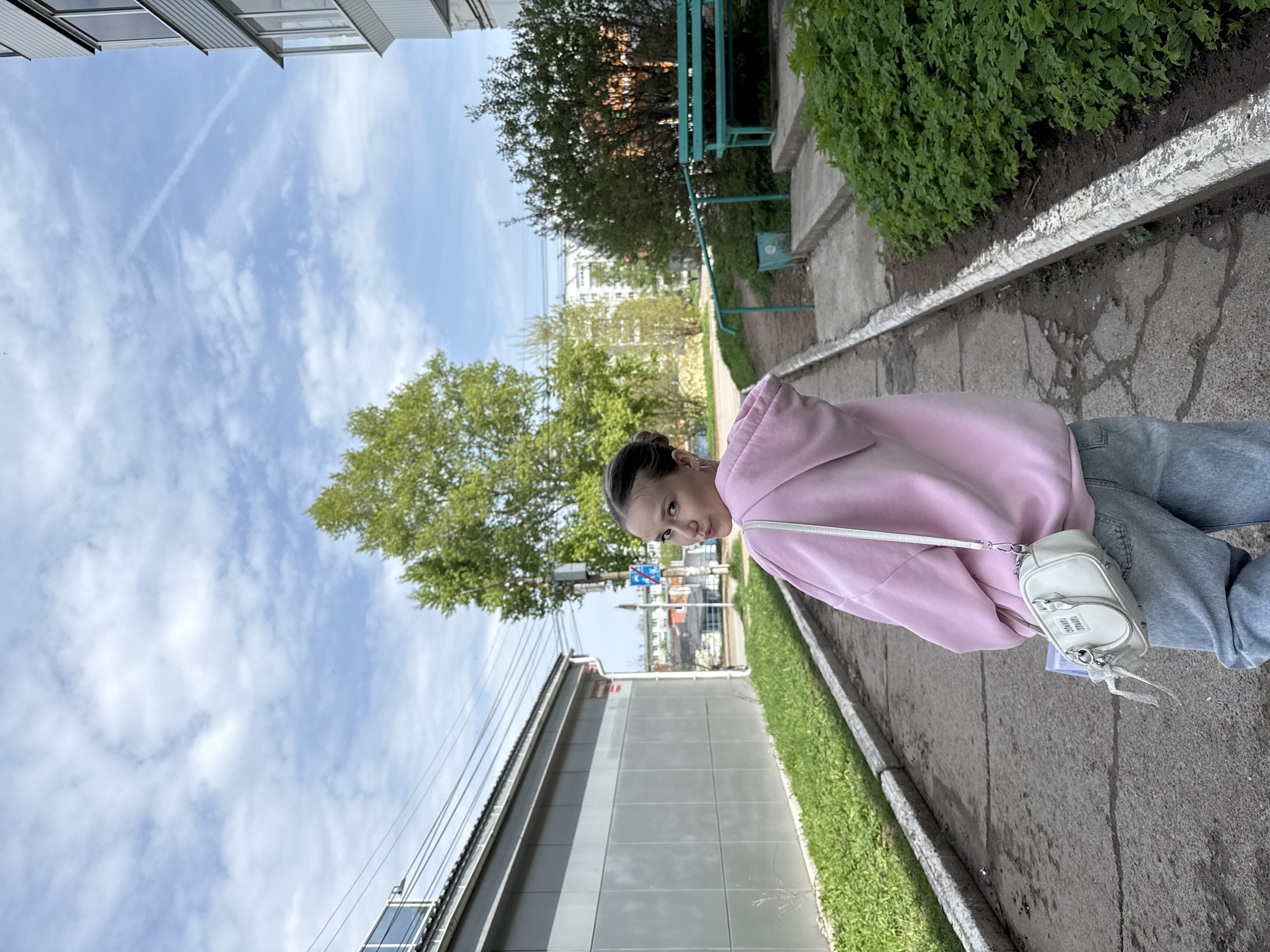A woman wearing a pink hoodie and blue jeans stands on a sidewalk in the city of Chaikovsky, Russia. She has a white purse slung over her shoulder. The sidewalk runs parallel to a building with a gray, corrugated metal facade. Across the sidewalk is a grassy area and some foliage. In the background, trees and other buildings can be seen, some appearing to be residential. Above, the sky is blue with scattered clouds.