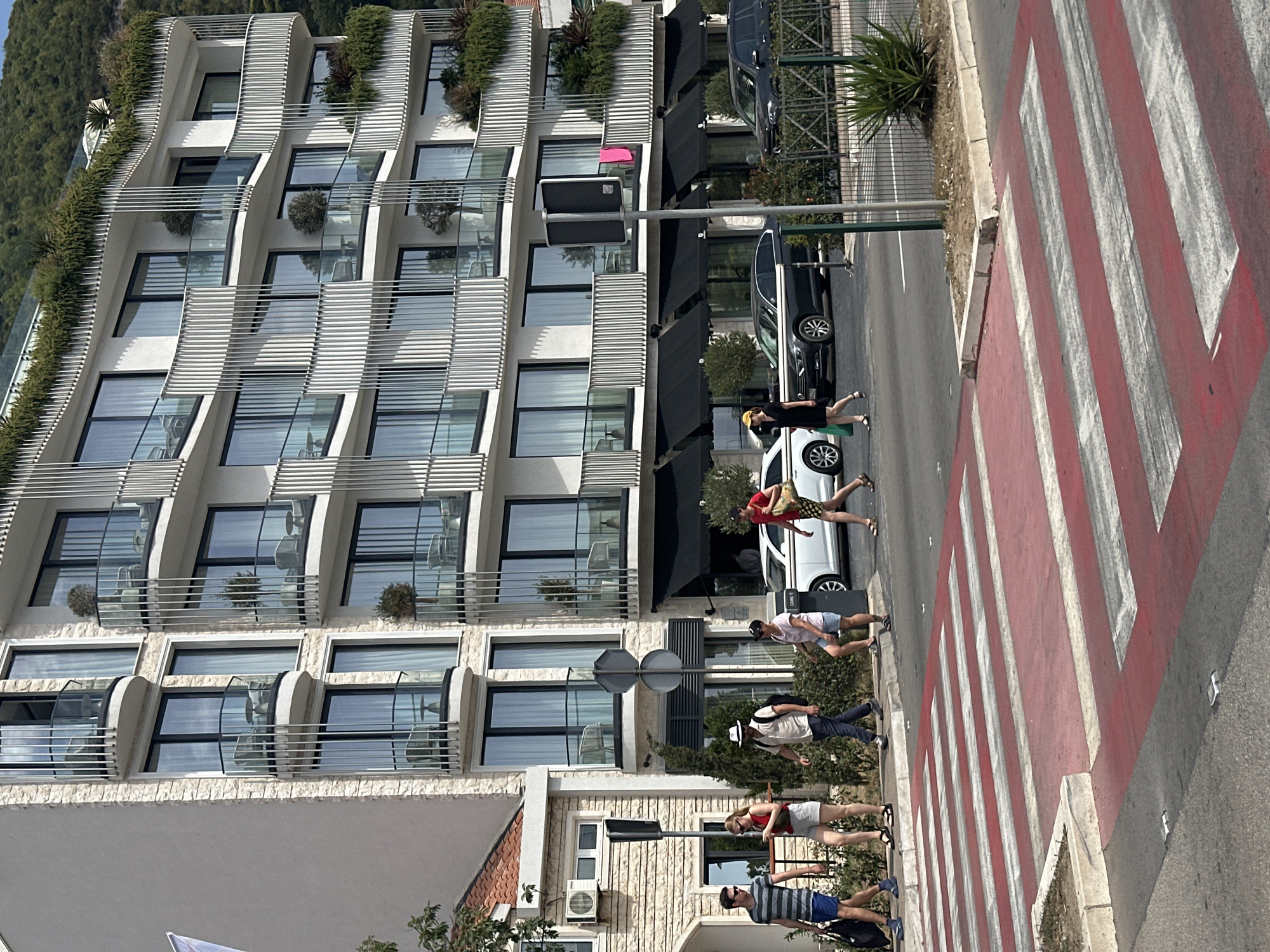 The image depicts a street scene in Budva, Montenegro, with a modern, multi-story building as the dominant backdrop. The building features numerous balconies with glass railings and vertical metal supports, with some balconies containing small trees and seating. The lower level of the building has black awnings extending over storefronts.
In the foreground, a red and white striped pedestrian crossing is visible. Several people are crossing the street on this zebra crossing, appearing to be tourists. From left to right, a man in a striped t-shirt and blue shorts walks with a black bag. Next to him, a woman in a red top and white shorts looks at her phone. A man in a white hat, cream-colored shirt, and dark pants follows. Another woman, wearing a pink t-shirt and denim shorts, walks with her hands in her pockets. Finally, a person in a red top and patterned shorts is crossing.
Cars are visible on the street. A white car is stopped near the crossing, and a dark-colored sedan is further down the road. A traffic light pole with two signs stands near the crossing.
The environment appears to be a sunny day, with the building casting a shadow across parts of the street. The overall impression is a typical urban streetscape with pedestrian activity and modern architecture.