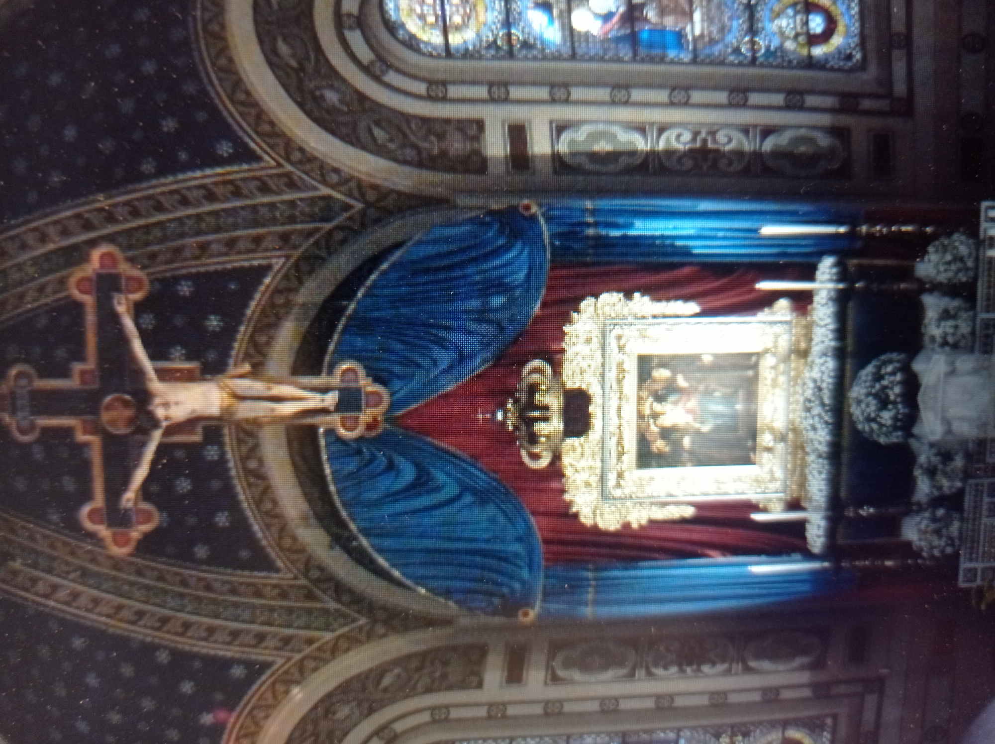 This is a photograph of the interior of the Church of the Ascension of the Lord in Mukachevo, Ukraine, focusing on the altar area. Dominating the upper portion of the image is a large crucifix with a figure of Jesus Christ. Below the crucifix, a framed religious icon depicting the Virgin Mary and Child is displayed, framed by ornate golden trim and topped with a crown. The icon is set against a backdrop of red and blue draped curtains. In front of the icon, an altar is visible, adorned with floral arrangements. Two tall white candles are placed on either side of the altar. The architectural style features arched ceilings and stained-glass windows on the right side of the frame.
