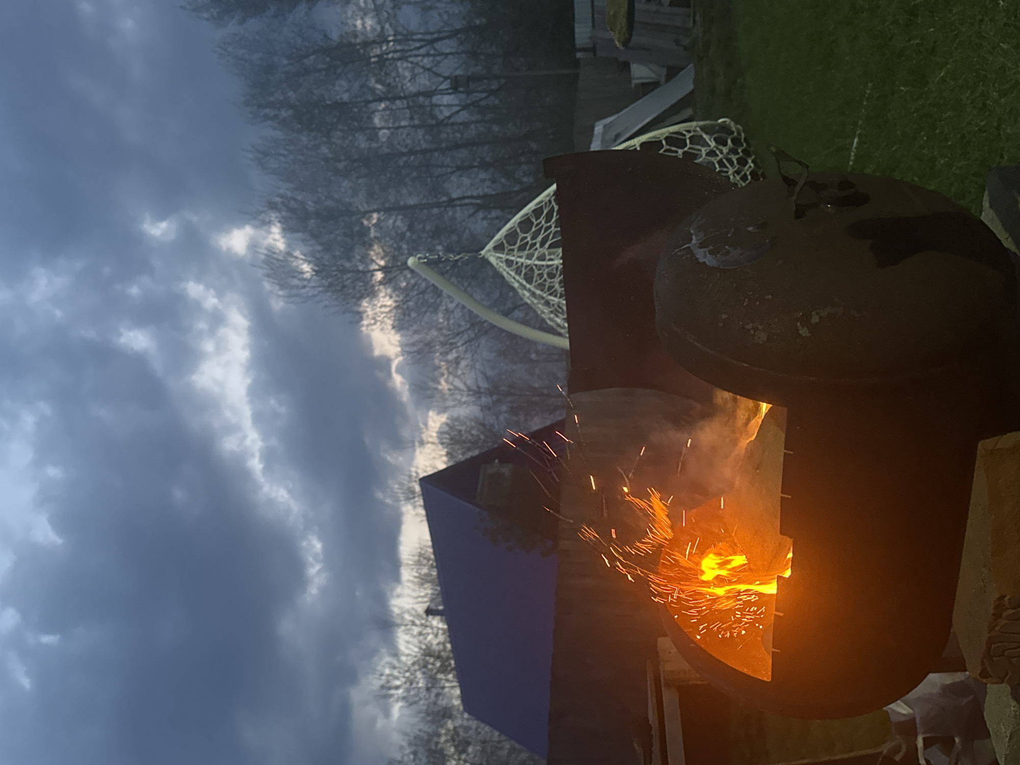 A sideways outdoor shot shows a lit fire pit in front of a cloudy sky and bare trees. The fire pit, located on a grassy lawn next to a play set with a climbing net and blue-sided playhouse, emits sparks and smoke. The photo was most likely taken at dusk or dawn. No people are visible. There is no definitive evidence to conclude that the image was taken in Chaikovsky, Russia.