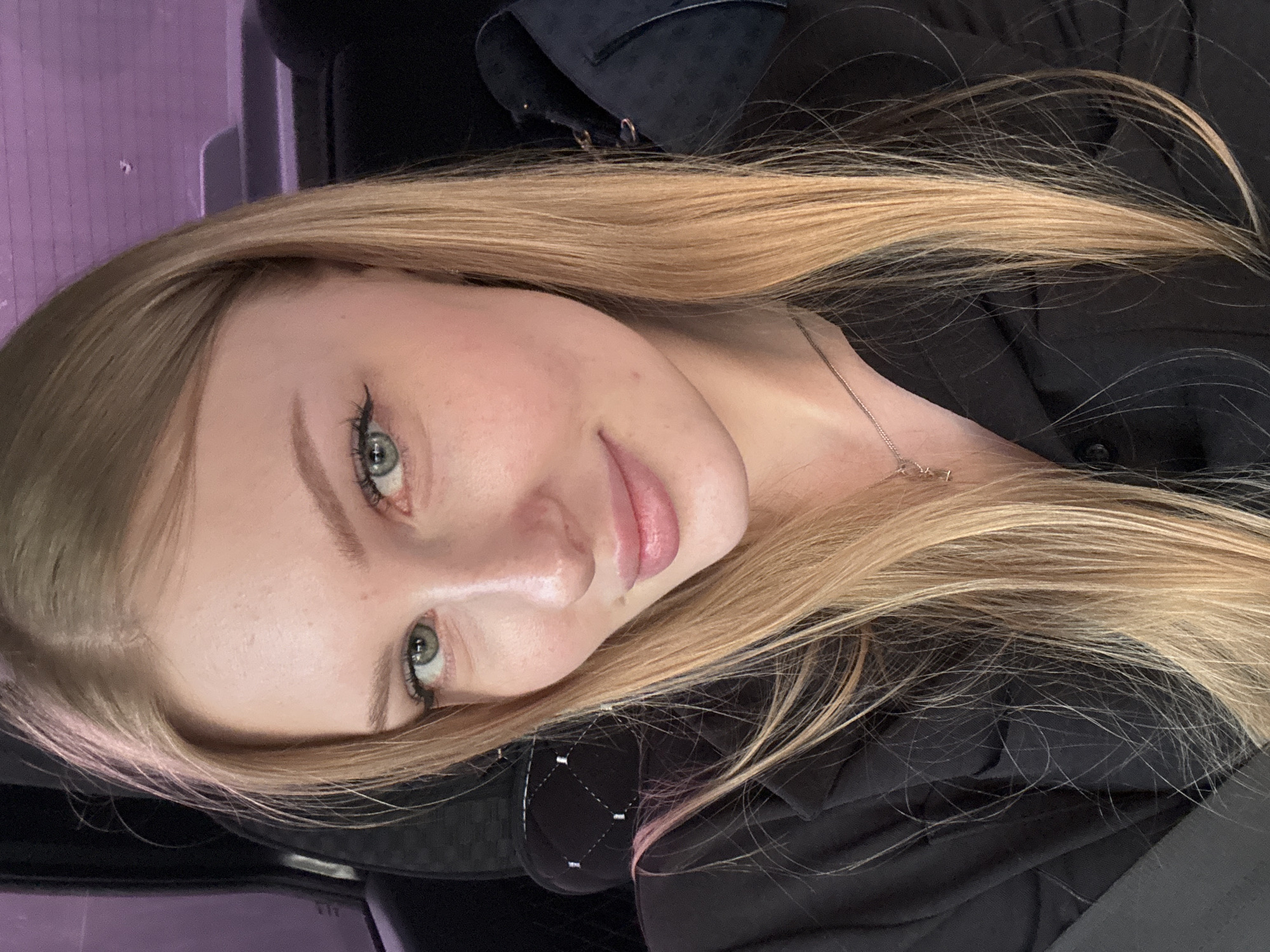 A selfie portrait of a light-skinned woman with long blonde hair and green eyes. She is wearing a black shirt and a thin necklace. The background includes a purple surface. The location is uncertain and cannot be verified.