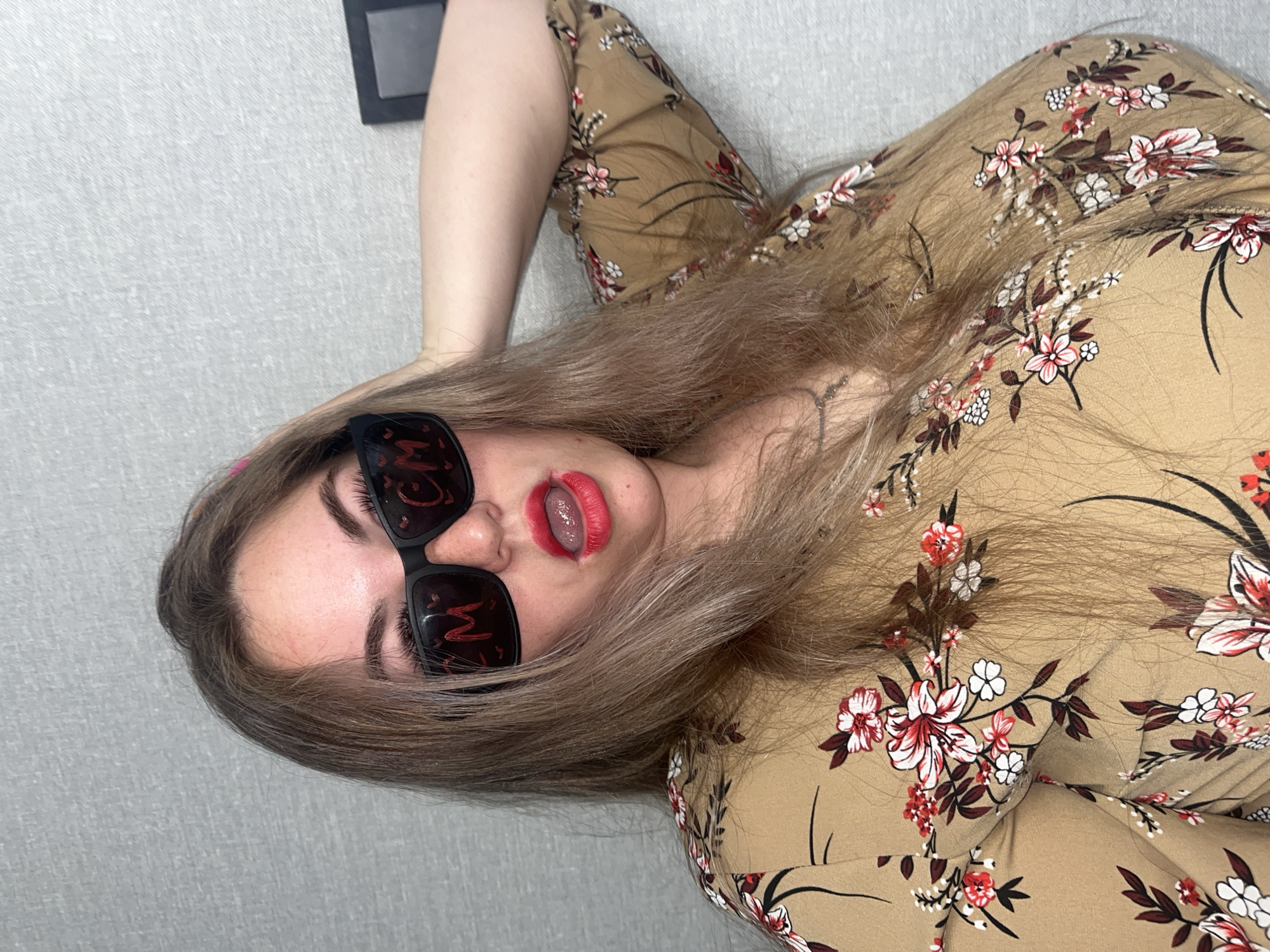 A person lies on a textured, light gray surface. They are wearing a beige floral print shirt and black sunglasses with red text reflected on the lenses. The person's hair is blonde and shoulder-length. Their mouth is open, revealing the tongue. In the upper left corner is a dark gray rectangular object. There is no indication of the location being Chaikovsky, Russia, in the image.