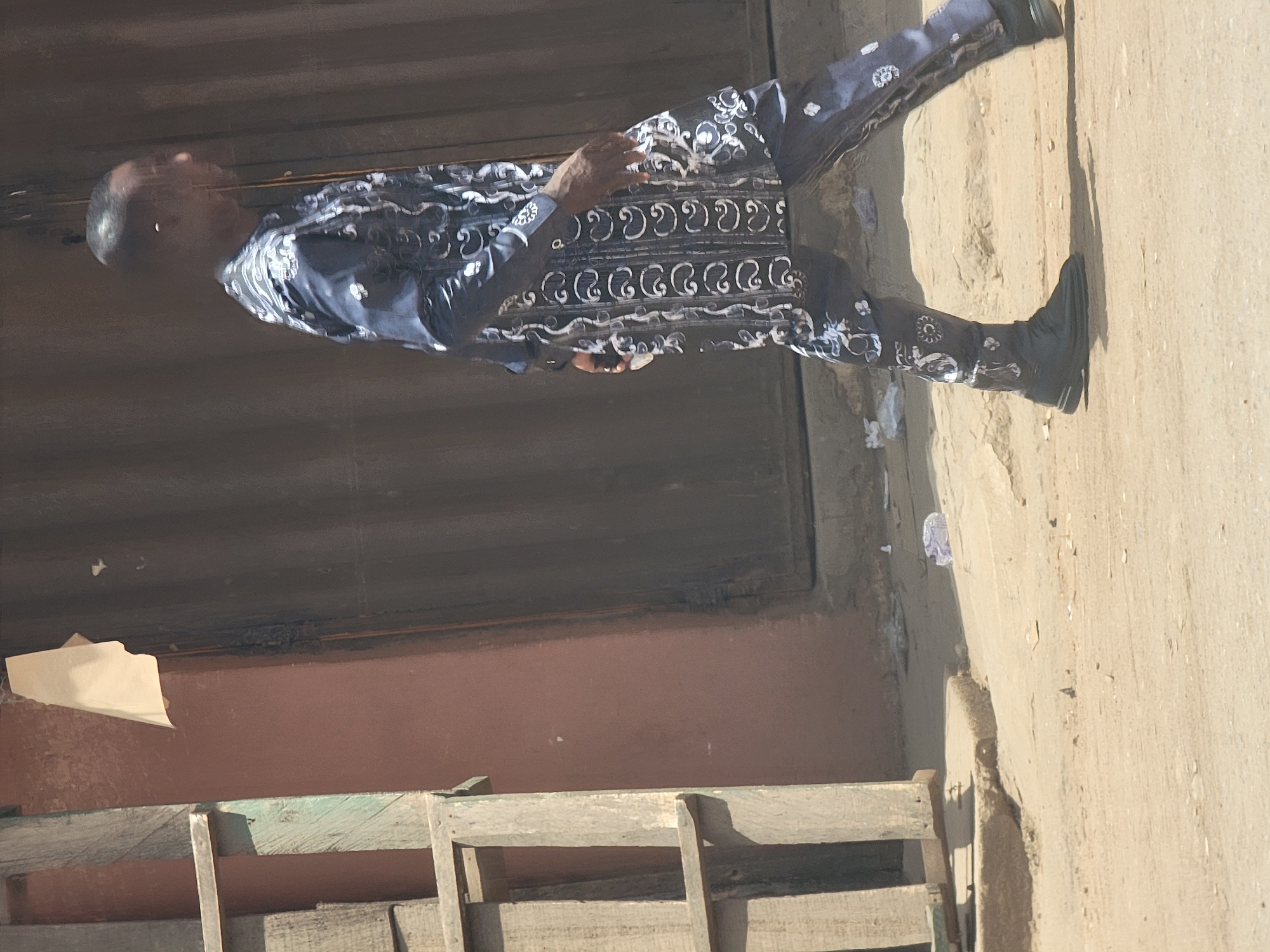 A man stands on a tan-colored surface next to a building in what could be a street scene in Maiburiji, Nigeria. The building behind him is painted a dull red and has a dark brown overhang. Several wooden chairs or crates are in front of the building. The man is wearing a dark blue suit with a repeating white pattern. He is also wearing black shoes. The overall image appears slightly tilted.