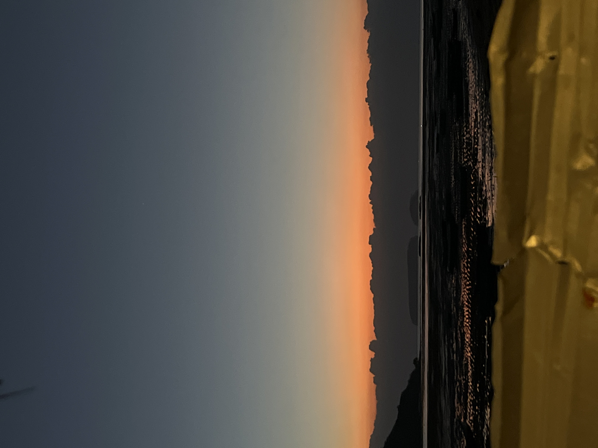 This is a vertical, eye-level, medium shot of a sunset over a beach in Kertasari, Indonesia. The sky transitions from a dark navy blue at the top to a vibrant orange band just above the horizon, with the last vestiges of sunlight reflecting off the wet sand. The foreground is dominated by the blurry, out-of-focus yellow surface of an unidentified object, possibly a beach umbrella or a piece of furniture. In the midground, the exposed beach is dark and textured, showing ripples and patterns from receding tide. Islands are visible in the distance, silhouetted against the colorful sky. The overall mood is tranquil and serene, characteristic of a peaceful evening by the sea.