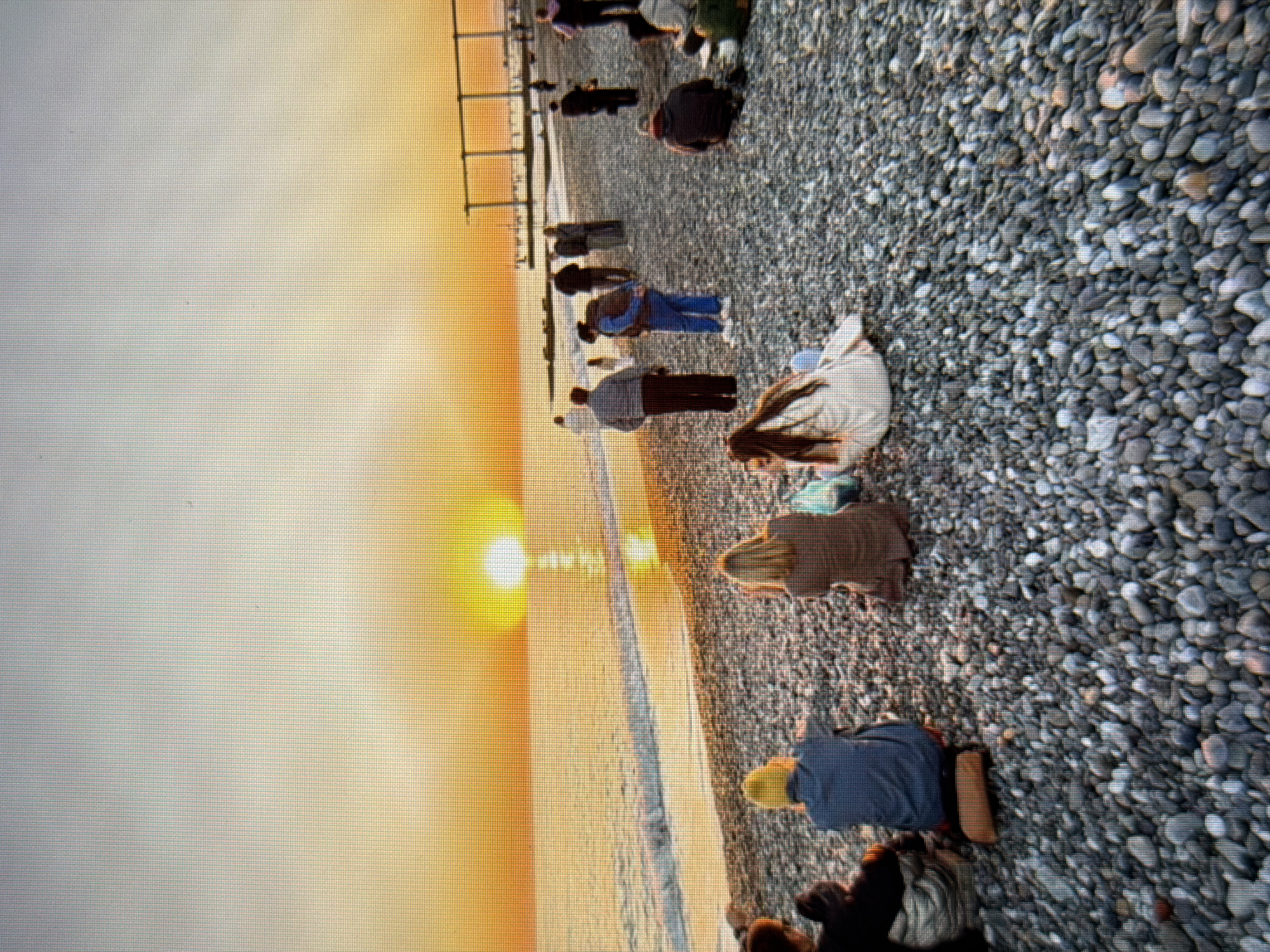 The image depicts a pebble beach in Sirius, Russia, during sunset. The sun is low on the horizon, casting a warm orange glow across the sky and reflecting on the sea. Several people are scattered across the beach, some sitting and facing the sea, others standing and walking. In the foreground, three individuals are seated, one wearing a yellow beanie, another in a brown jacket, and a third in a light-colored coat. Further back, more people are visible, with a few individuals walking along the water's edge and a structure resembling a pier or promenade in the distance. The scene conveys a sense of calm and relaxation as people gather to enjoy the sunset.