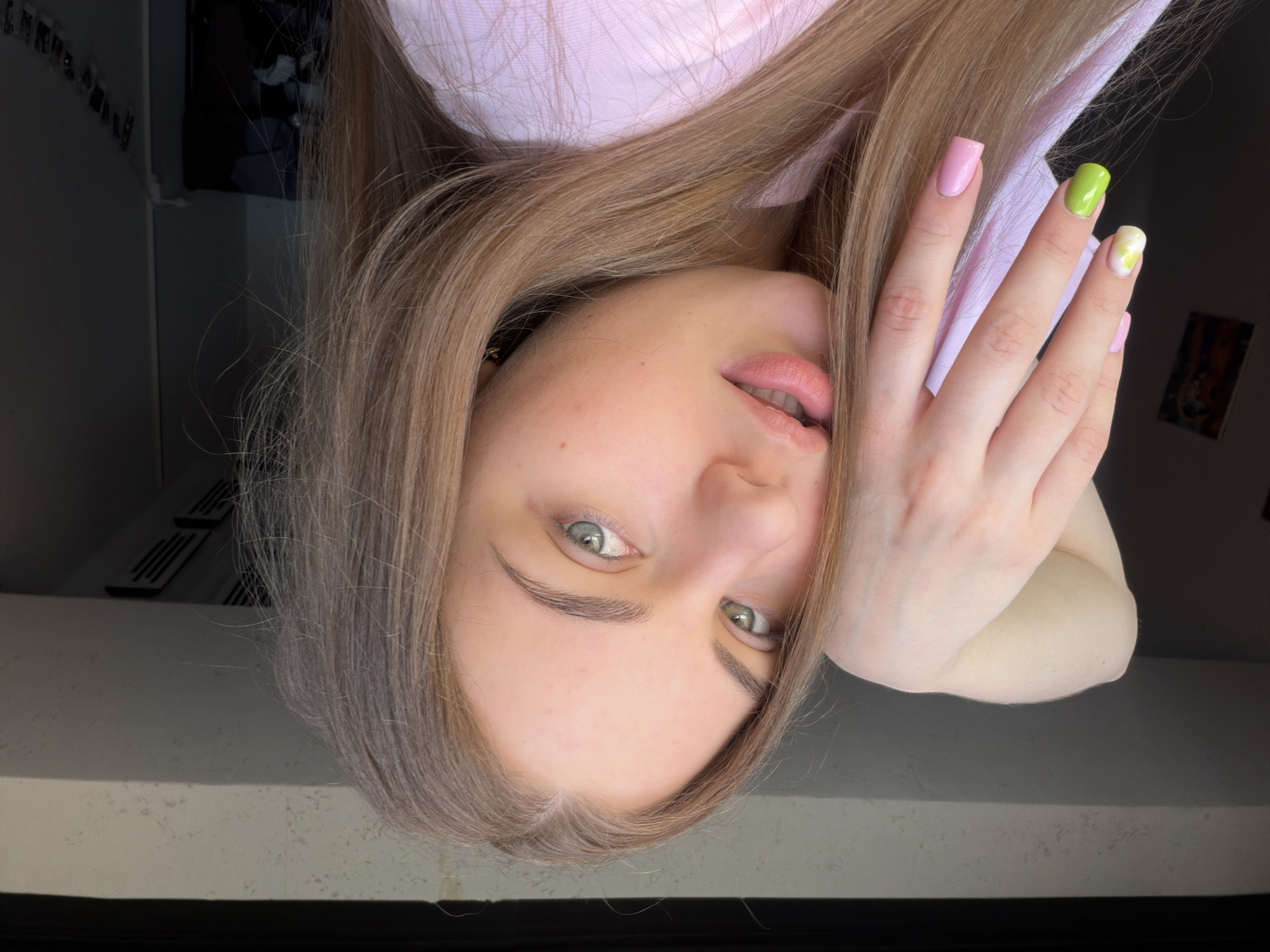 A close-up, head-and-shoulders shot captures a young woman with light brown hair and blue-green eyes looking directly at the camera. Her left hand is raised, partially obscuring her face, with nails painted in pink and green with a swirled design on one nail. She is wearing a light pink, collared shirt. The background is indistinct and dark, with a sliver of what appears to be a window frame visible at the top. The lighting suggests an indoor setting, possibly near a window. There is no discernible indication of the specific location within the city of Dnipro, Ukraine, or any urban environmental details.