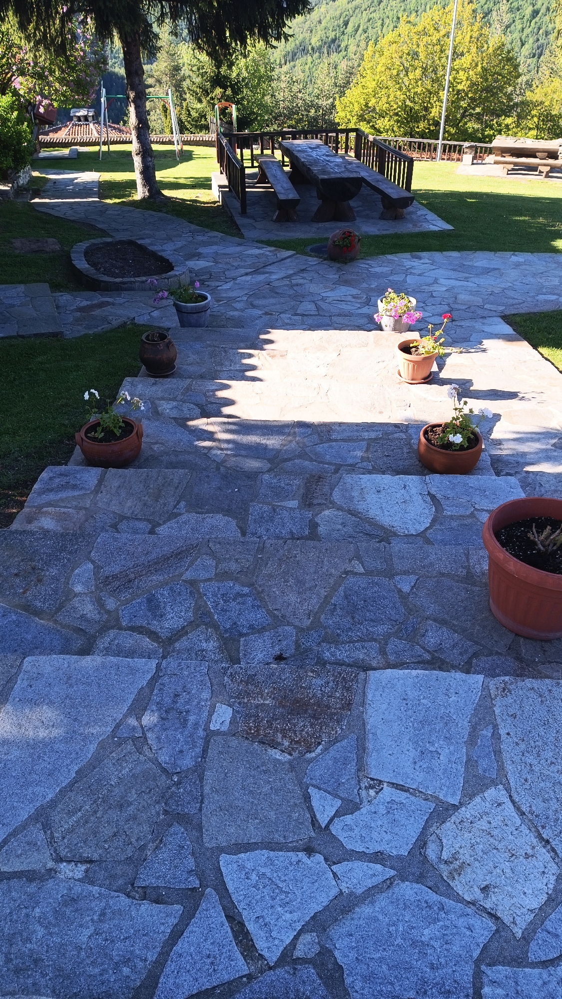 The image depicts an outdoor scene in Vrata, Bulgaria. A stone pathway descends a set of steps made of irregularly shaped blue-gray stones. Potted plants are placed on the steps and at the top of the staircase, adding touches of color. The area at the top of the stairs is paved with similar stones, leading to a picnic table and benches. Beyond the patio, a grassy area extends towards trees and a forested hillside. Sunlight is visible, with shadows cast across the scene, suggesting it is daytime. There are no people visible in the image. The environment is tranquil and natural, reflecting a rural or semi-rural setting.