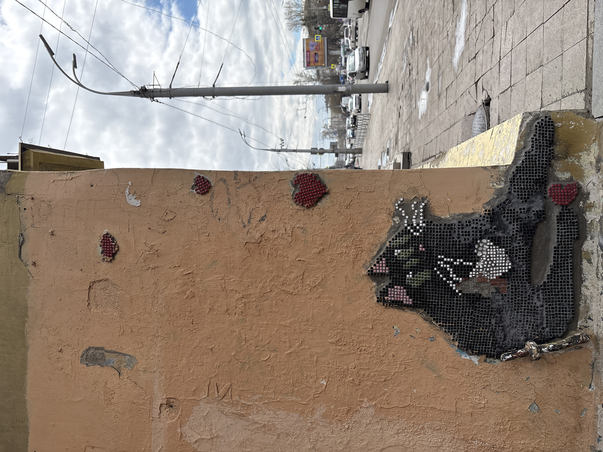 The image shows a section of a dilapidated wall in Irkutsk, Russia. The wall is painted a faded peach color with peeling paint. There are three heart-shaped mosaics made of red tiles adhered to the wall. To the right, a larger, irregularly shaped mosaic is embedded in the wall, depicting a black cat among pink and white flowers. The mosaic is partially damaged, revealing the mesh backing and the wall beneath. A metal pipe protrudes from the wall near the mosaic.
In the background, the image depicts a street scene with cars, buses, and utility poles under a cloudy sky. The pavement appears to be made of paving stones. A building with signage is visible across the street.