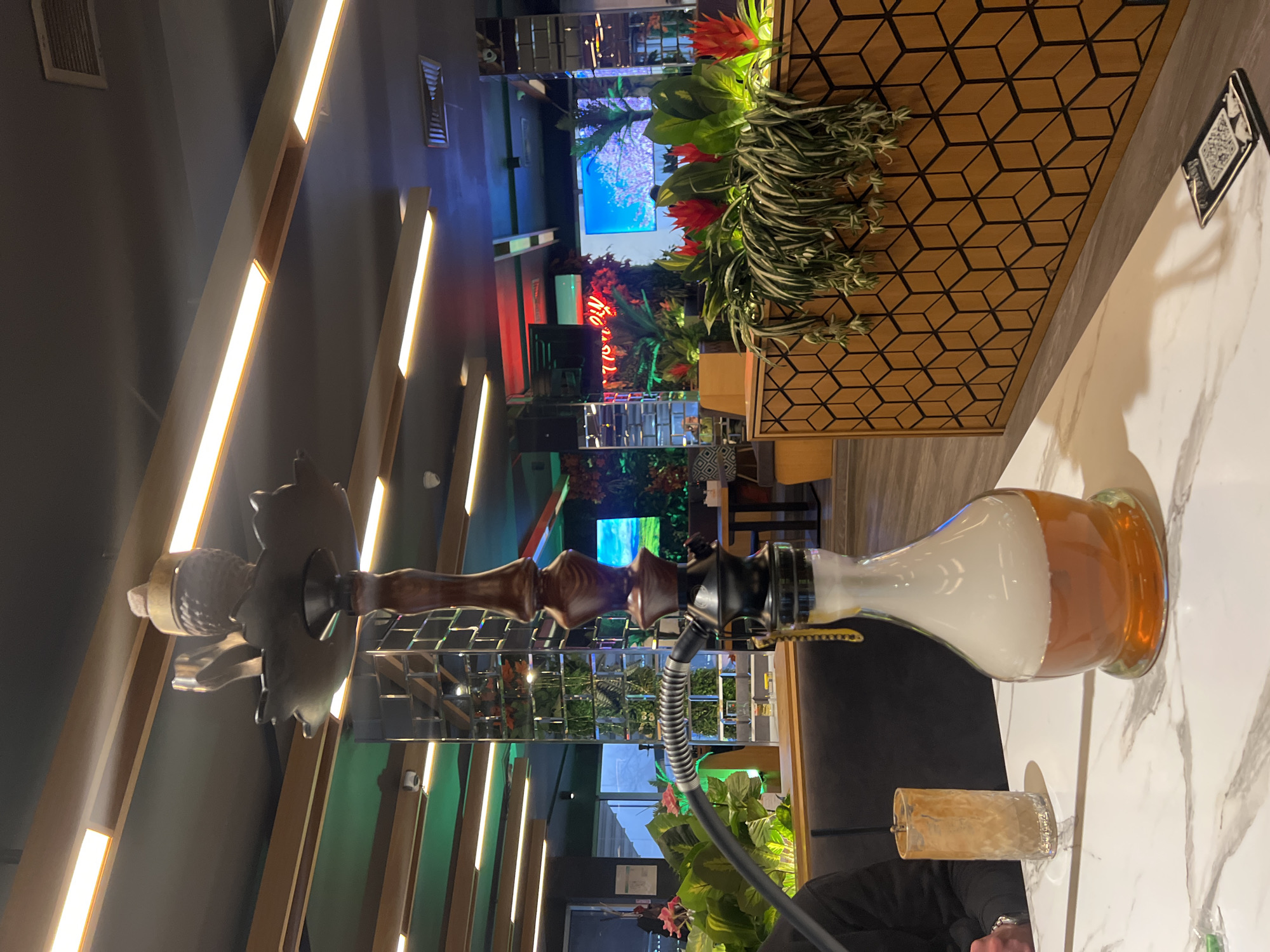 The image depicts the interior of a modern lounge or cafe located in Sofia, Bulgaria, characterized by contemporary design and ambient lighting. In the foreground, a prominent shisha (hookah) stands on a white marble-patterned table. The shisha features a dark brown wooden stem, black metallic components, and a clear glass base containing an amber-colored liquid with white vapor concentrated at the top. A black corrugated hose extends from the shisha towards the left.
To the left of the shisha, a textured clear glass with a residual amber liquid is visible. A person's left forearm and hand, wearing a metallic wristwatch and a black long-sleeved garment, are positioned near the glass and shisha hose. On the right side of the table, a small black rectangular object with a QR code is present.
The background features dark grey upholstered booth seating. A wooden divider with a geometric cube pattern supports a collection of green plants, some bearing red flowers, and trailing foliage. Further back, mirrored pillars composed of square tiles reflect elements of the interior. Multiple digital displays are visible, one showing a natural landscape scene with pink-blossomed trees, and another, partially obscured, displaying red neon text. The ceiling is dark grey, equipped with recessed linear light fixtures providing warm illumination, and includes visible air vents and small white dome security cameras. Green and red accent lighting contributes to the atmosphere. A large opening on the far left reveals an exterior or an adjacent section of the building, possibly including stairs.