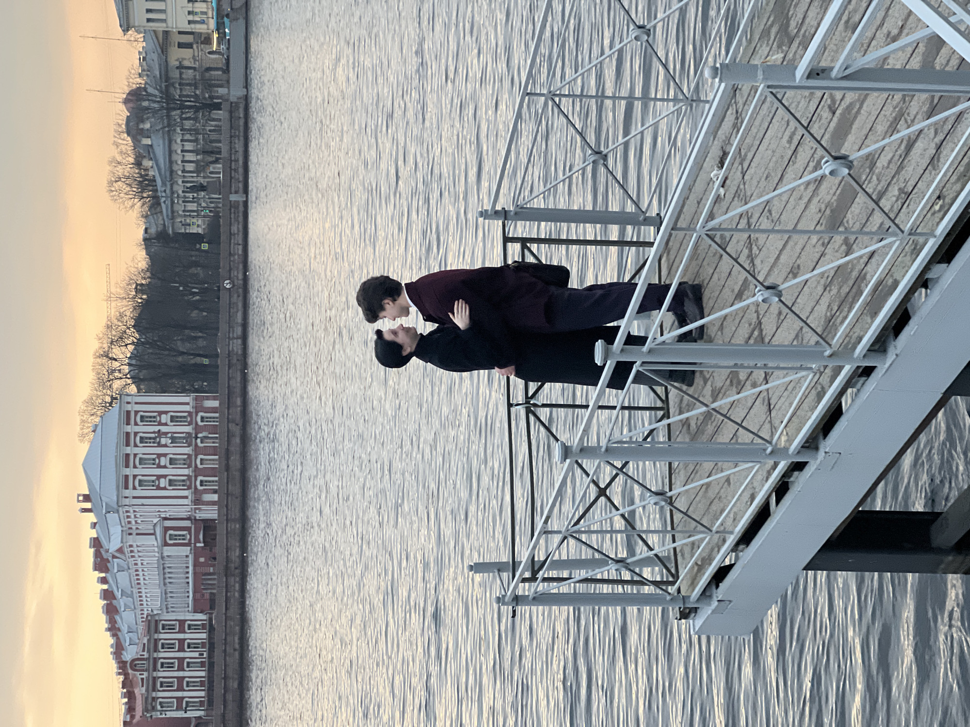 Two males stand embraced on a bridge over water. One, dressed in black, wears a hat. The other, in a dark burgundy overcoat, leans into the first. Behind them, across the water, lies a building with a red and white facade. The sunset paints the sky with soft hues. The location is Saint Petersburg, Russia.