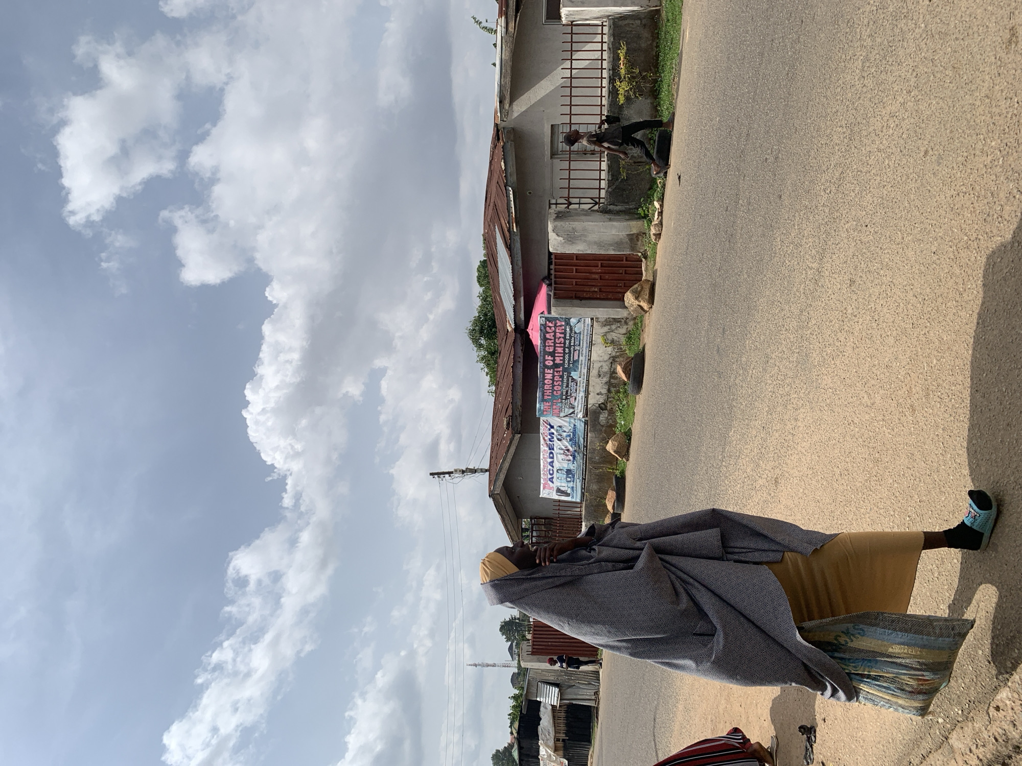 A middle-aged woman stands on an asphalt road in Jos, Nigeria. She wears a yellow top, carries a striped bag, and is draped in a gray shawl. A building with a metal fence and signs for "Throne of Grace Gospel Ministry" is visible. Clouds fill the sky. A person walks along the roadside near the building. Other buildings line the street further in the distance.