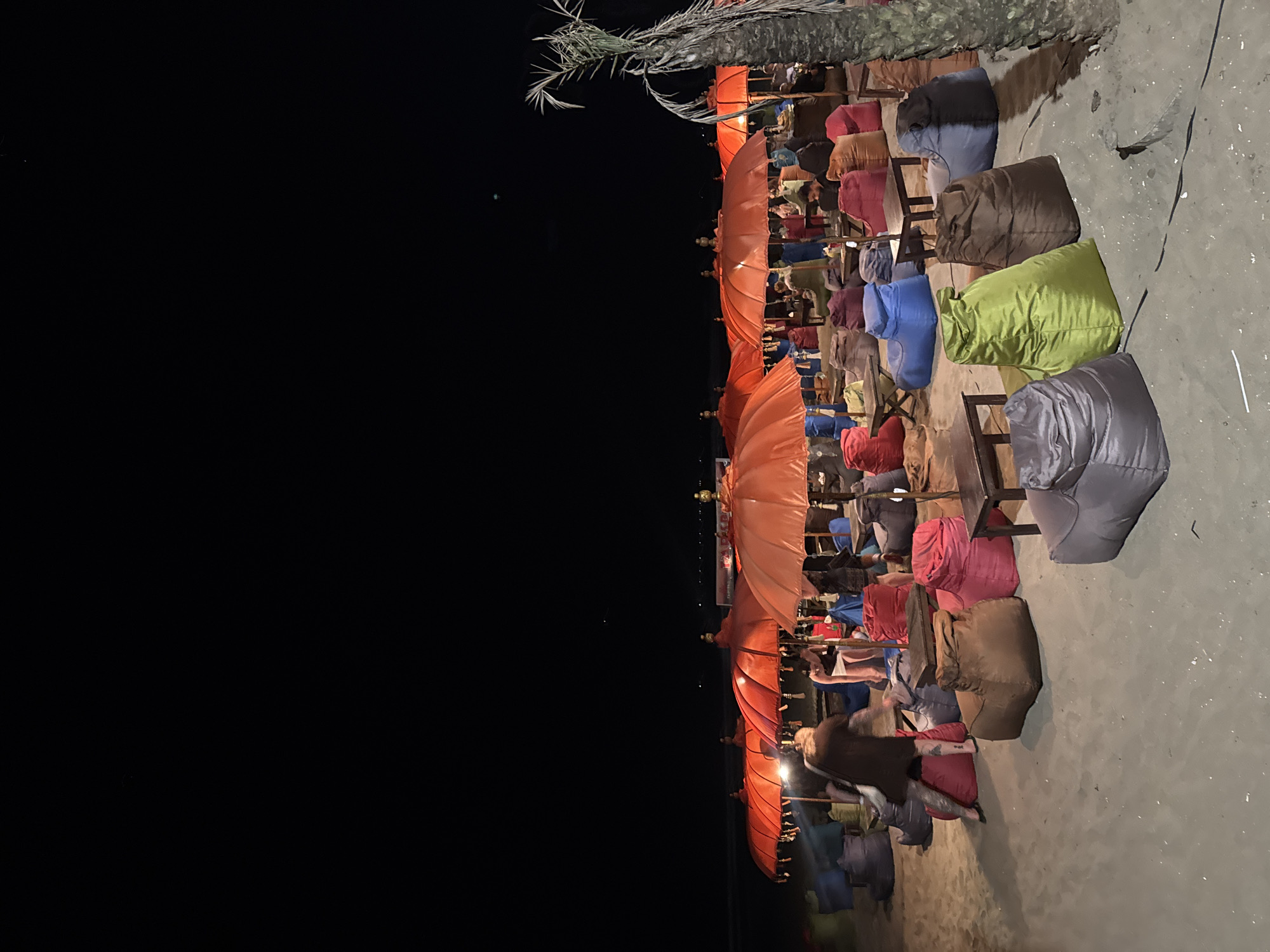 The image depicts a nighttime scene on a sandy beach in Kuta, Indonesia, likely a beach bar or restaurant. Several large, brightly colored orange umbrellas are set up, providing shade and ambiance. In front of the umbrellas, numerous beanbag chairs in various colors such as blue, red, brown, green, and gray are arranged on the sand, with small wooden tables interspersed among them. A few people are visible, some seated on the beanbags and others standing or walking. One person in the foreground is wearing a dark dress and appears to be walking. The background shows the dark expanse of the night sky and the faint lights of what could be distant buildings or boats. A palm tree is visible on the right side of the image. The overall atmosphere is relaxed and social, suggesting a popular spot for evening relaxation on the beach.