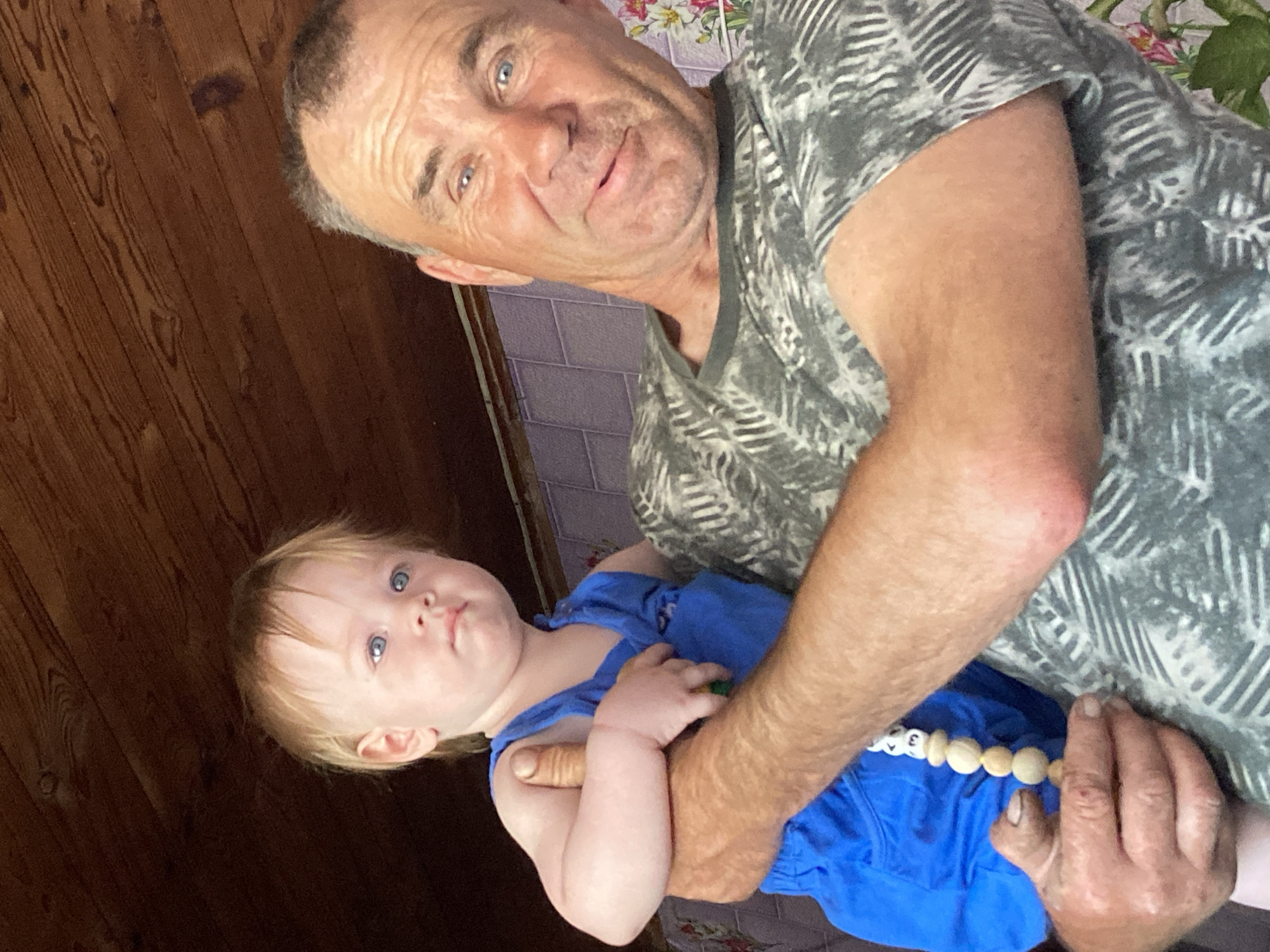 In this close-up, waist-up indoor photograph, an older man with graying hair and blue eyes holds a young child. The man, wearing a grey t-shirt with a black and white leaf pattern, is looking towards the camera with a slight smile. His right arm is around the child, whose left arm is resting on the man's chest. The child, with blond hair and blue eyes, is wearing a blue sleeveless outfit and has a string of wooden beads hanging from their hand. The background consists of a wooden ceiling and a tiled wall with a floral pattern. The scene appears to be in a domestic setting.