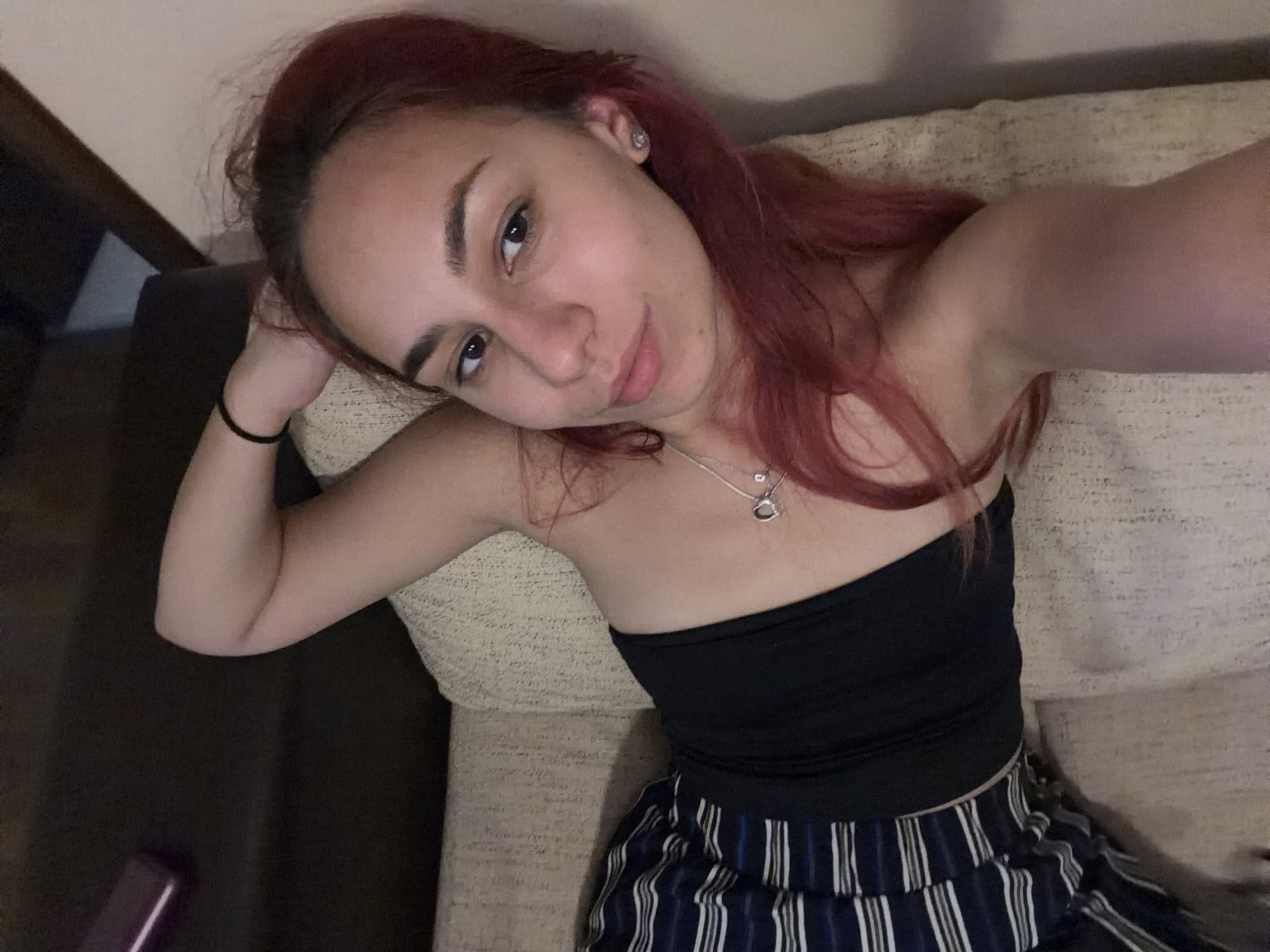 A young woman with auburn hair is lying on a couch, looking at the camera. She is wearing a black tank top and striped pants. Her left arm is bent behind her head, and she is wearing a silver necklace with a heart-shaped pendant. The couch appears to be in a home setting. The image does not contain any information to indicate it was taken in the city of Nesebar, Bulgaria.
