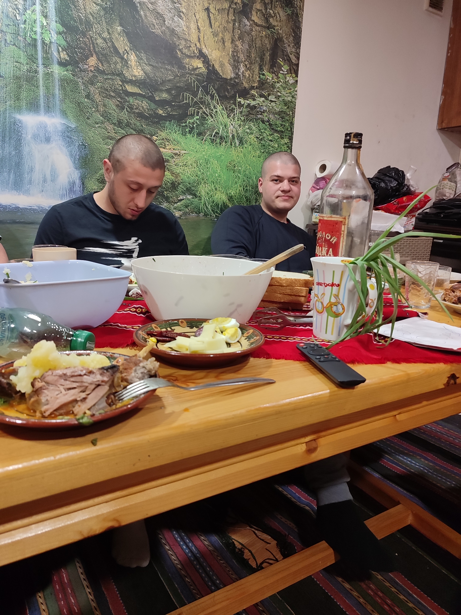 Two men are seated at a wooden table laden with food, including meat, potatoes, eggs, bread, and salad. A bottle of what appears to be alcohol and a mug are also on the table. The table is covered with a red and patterned tablecloth. A remote control rests on the table. One man is looking down, while the other looks toward the camera. The background features a wall covering depicting a waterfall and forest scene. A cabinet is visible on the right side of the image. The location is not specified, nor is the city of Varshilo, Bulgaria mentioned in the provided image. It would be impossible to tell the image location with the provided information.
