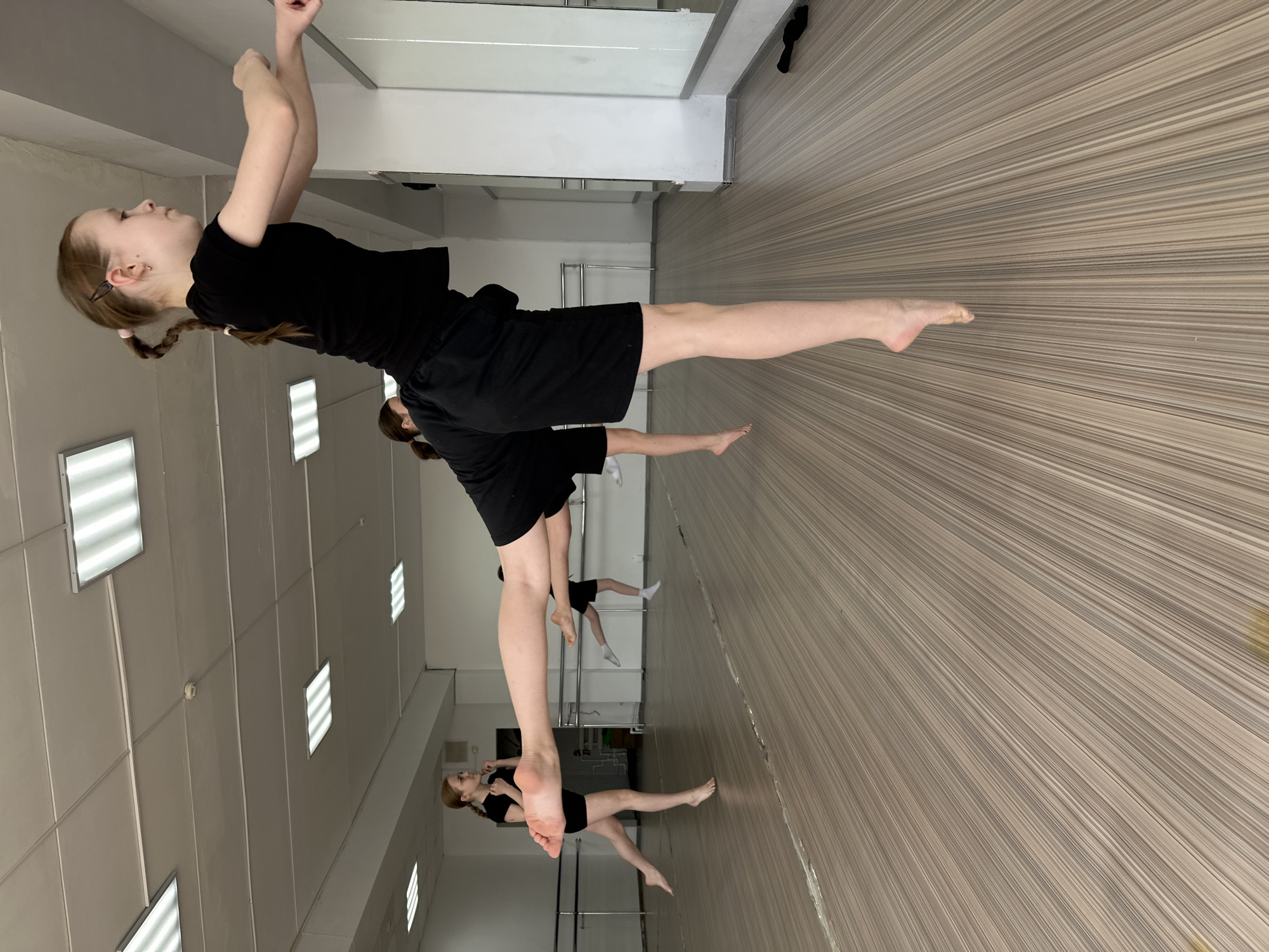 The image shows a group of young female dancers in a dance studio, likely in Votkinsk, Russia, given the information provided. The dancers are dressed in black shorts and shirts, and they are performing a synchronized dance routine. They appear to be barefoot. The studio has a wooden floor and a wall of mirrors. There are fluorescent lights on the ceiling. The room seems spacious.