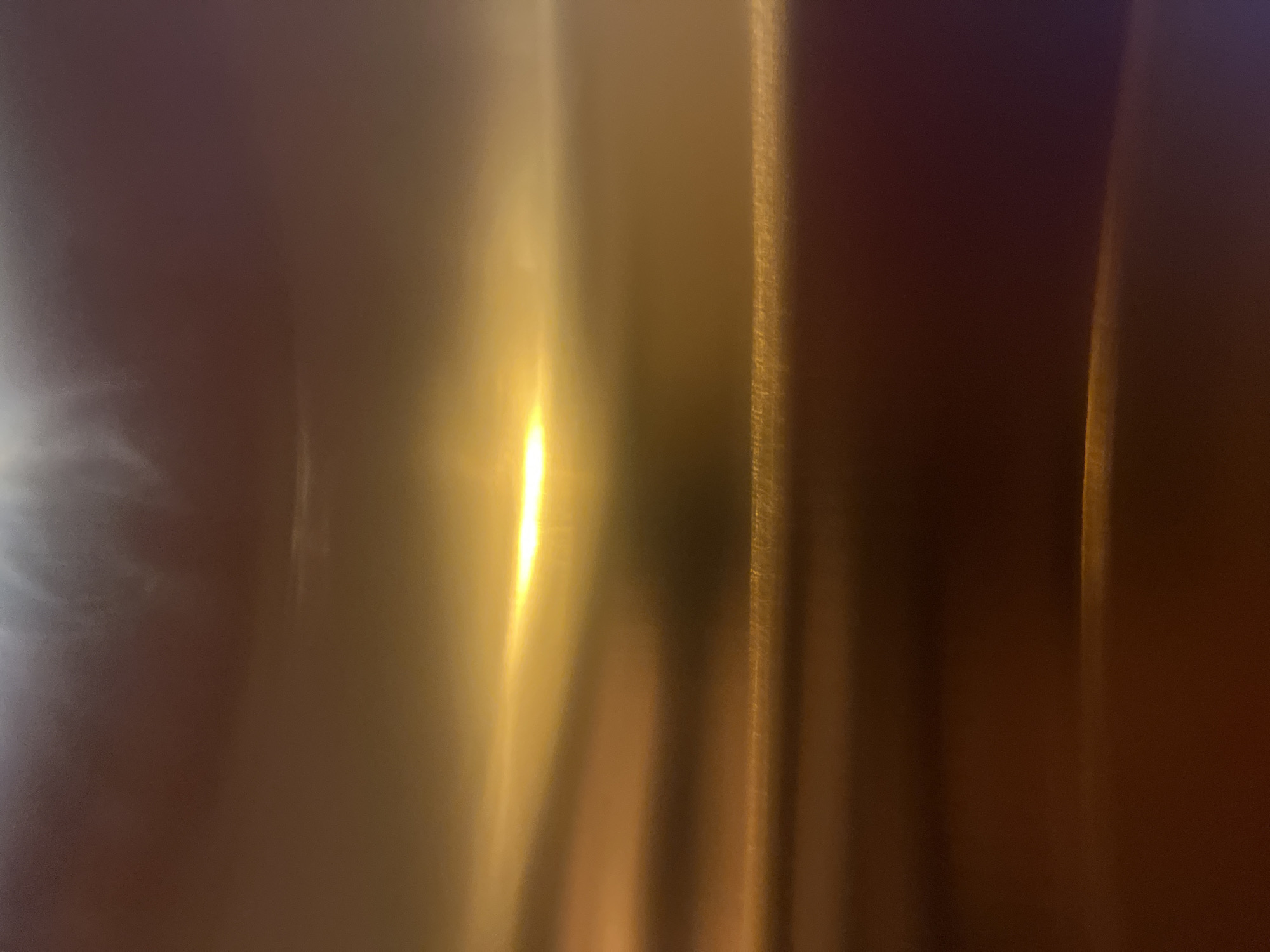The image depicts an abstract, out-of-focus scene dominated by warm tones of orange, yellow, and brown, with hints of purple in the lower right corner. A bright, horizontal band of yellow light cuts across the middle of the frame, suggesting a strong light source. The textures are soft and blurred, giving the impression of movement or a reflective surface. There are no discernible people, subjects, objects, or actions clearly identifiable. The image does not provide any specific visual cues that would allow for the determination of its location within the city of Boja, Indonesia, or any other specific location.