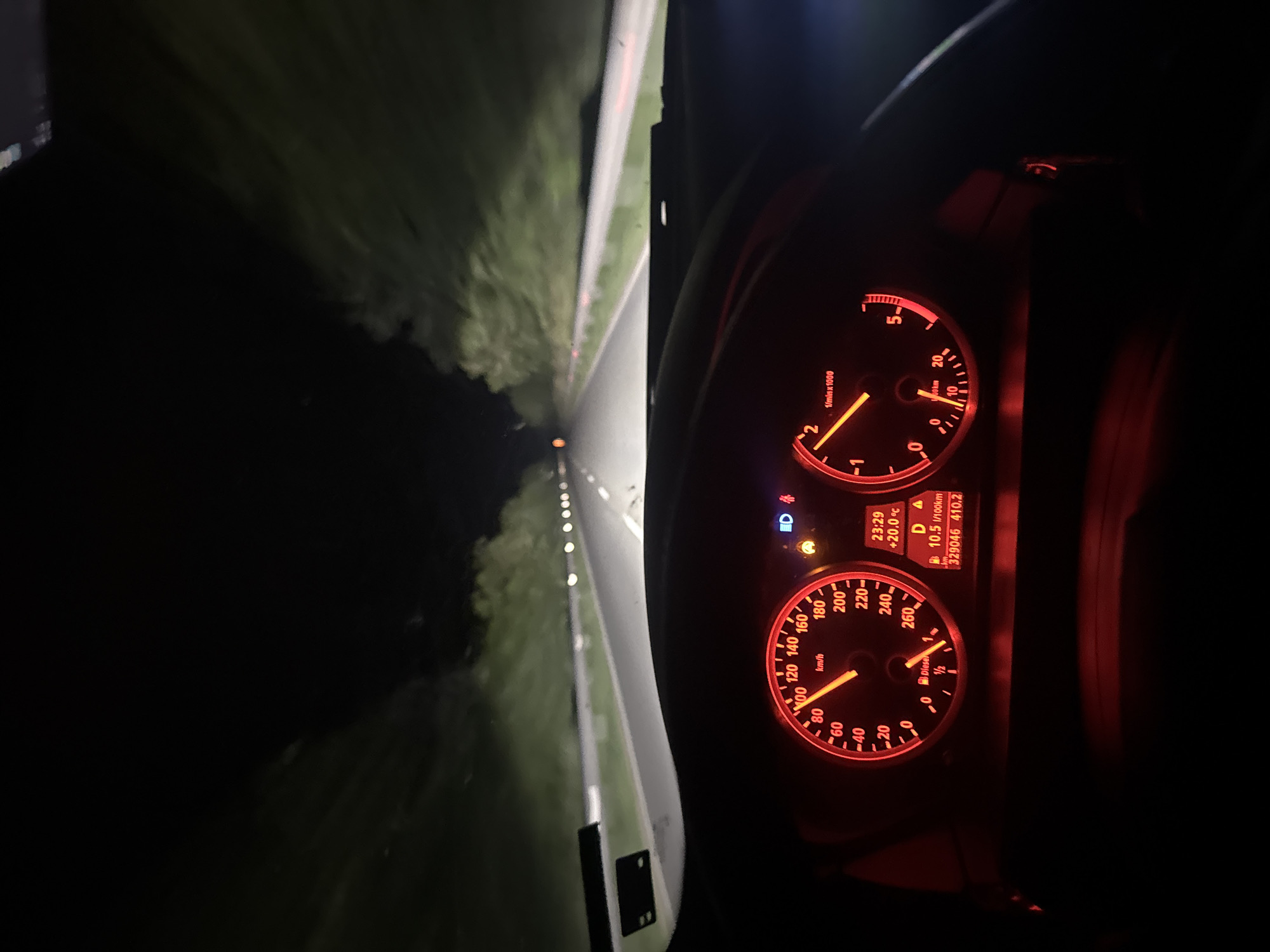 A nighttime view from inside a car driving on a road. The dashboard gauges are illuminated, showing a speed of approximately 120 km/h and an engine speed around 2000 RPM. The road is flanked by vegetation on either side, lit by the car's headlights and punctuated by evenly spaced lights. A location in the city of Stara Zagora, Bulgaria is not discernible from the image.