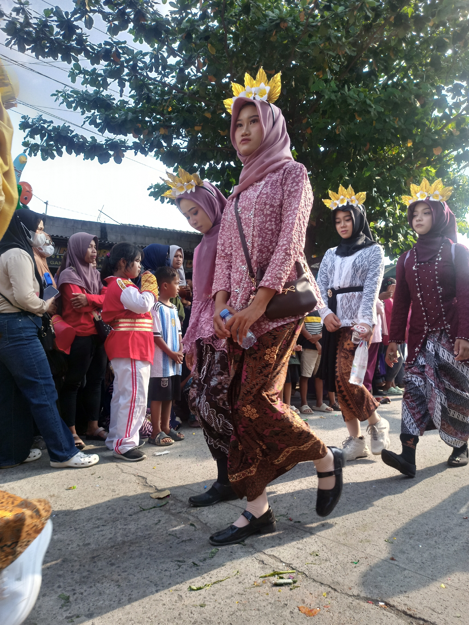 The image depicts a daytime street scene in Saren, Indonesia, likely during a cultural event or parade. Several women, some wearing traditional Indonesian attire including batik skirts and headscarves, are walking forward. A few of them are adorned with elaborate golden headpieces featuring floral motifs. One woman in the foreground wears a pink patterned top and a batik skirt, with a brown crossbody bag. Another woman to her left wears a similar pink headscarf and patterned top. To the right, a woman in a dark maroon outfit and headscarf also walks forward, with a patterned skirt. Further back, other individuals, including children, are visible in the background, some appearing to be spectators. The ground is paved, and overhead, lush green tree foliage casts shadows. Scattered on the ground are small pieces of debris, possibly from a celebration. The overall atmosphere suggests a festive gathering.