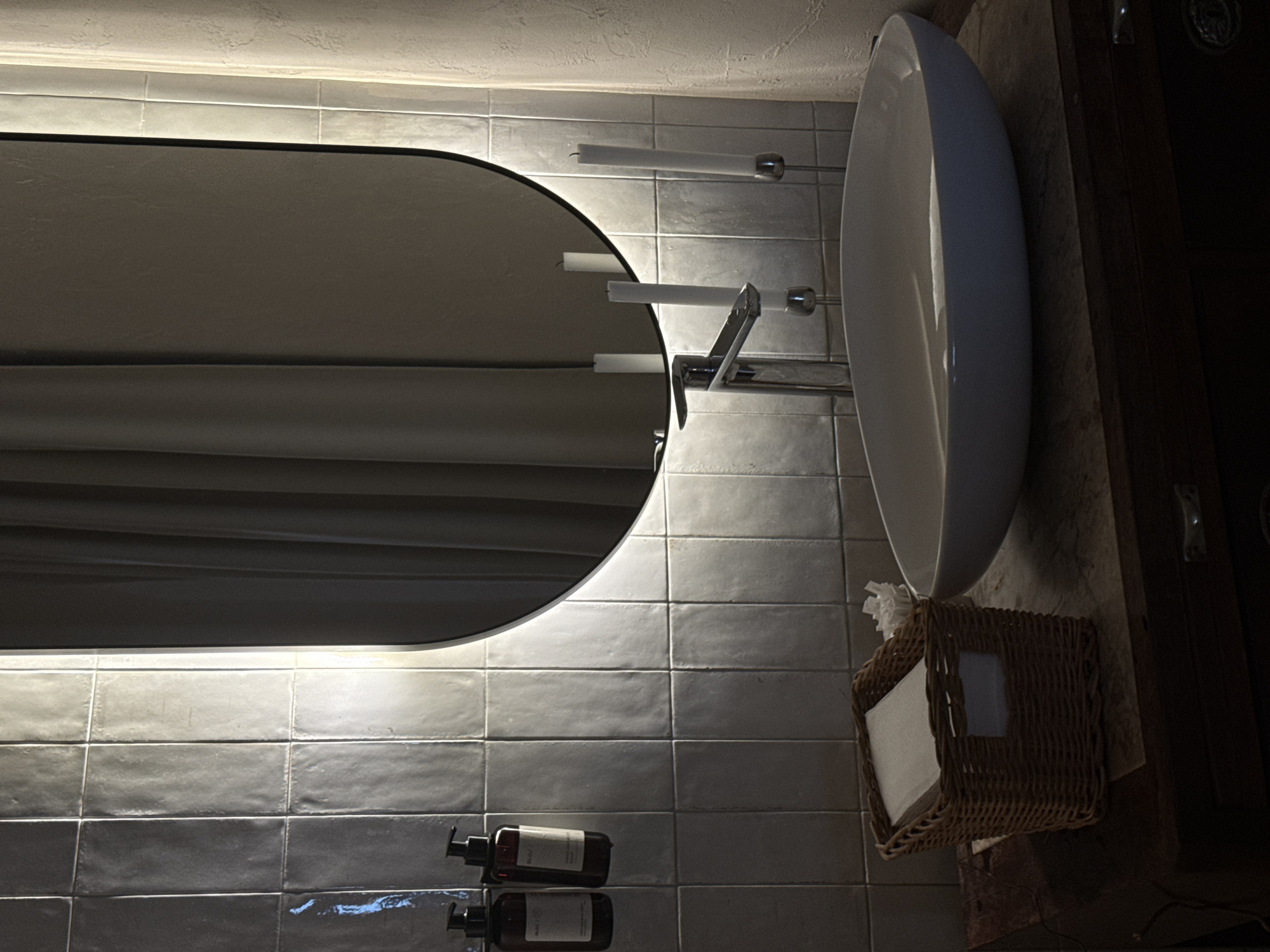 The image shows a bathroom interior. A large, vertically oriented oval mirror with a light strip is mounted on a tiled wall. The mirror reflects a curtain or similar fabric. A modern, white oval sink is positioned next to the mirror. Above the sink are two towel bars. A chrome faucet is mounted on the tile wall, extending over the sink. Below the mirror, on the tiled surface, are two pump bottles. A woven basket containing tissues is situated next to the sink, near a dark wooden cabinet or wall panel. The tiles on the wall are rectangular and appear to be gray. The lighting in the bathroom is dim. There are no people visible in the image. The location of the bathroom in the city of Belgrade, Serbia, is not determinable from the image.