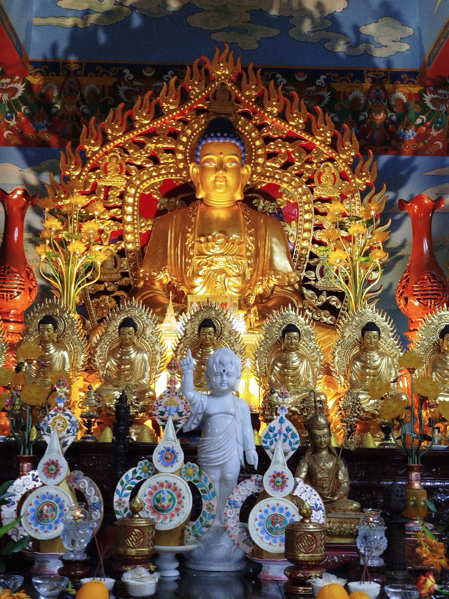 The image depicts an altar within a Buddhist temple, likely located in Da Lat, Vietnam, given the ornate style common in Vietnamese Buddhist iconography. At the center, a large, golden Buddha statue sits enthroned, adorned with a radiant aureole. Flanking this central figure are several smaller golden Buddha statues, arranged in rows and appearing to be in various meditative postures. In the foreground, a white marble statue of a young boy, possibly representing a celestial being or a child Buddha, stands with one hand raised. The altar is decorated with vibrant floral arrangements, golden ornamental pieces resembling lotus flowers, and intricately designed Buddhist symbols, including prayer wheels and mandalas. Two large, orange vases with ornate carvings are positioned on either side of the main Buddha statue. The background features a richly decorated mural, likely depicting mythological or religious scenes, characterized by dragons and celestial clouds. The overall atmosphere is one of reverence and spiritual devotion.