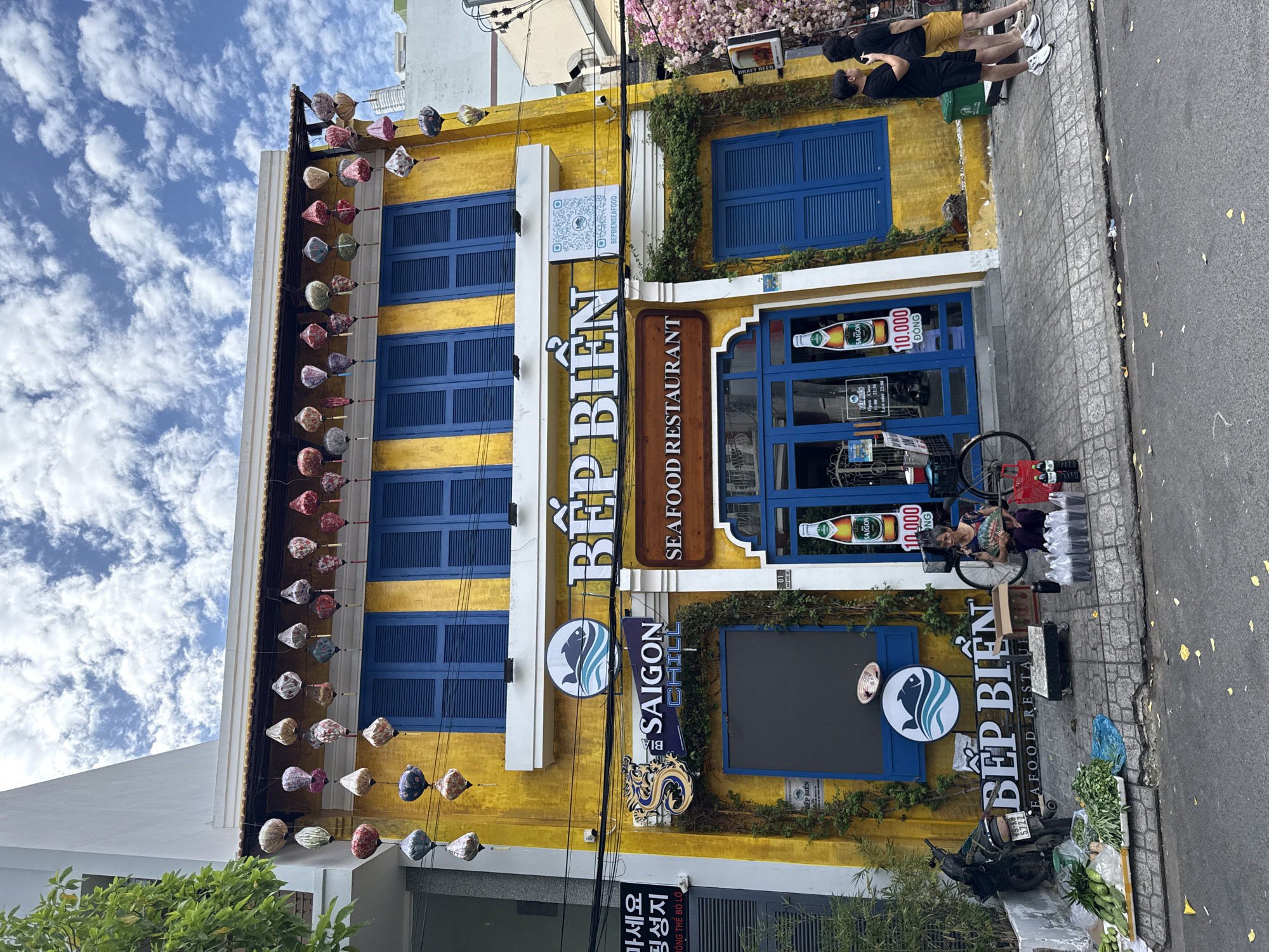 The image depicts the exterior of a yellow building with blue shutters in Nha Trang, Vietnam, identified as "BẾP BIỂN SEAFOOD RESTAURANT". The building's facade is adorned with numerous colorful paper lanterns hanging from an overhang. In front of the restaurant, a street vendor is seated on the sidewalk, surrounded by produce and bags. Two individuals stand further down the sidewalk, facing the building. The scene is set on a street with paved sidewalks and a road, with trees and other buildings visible in the background.