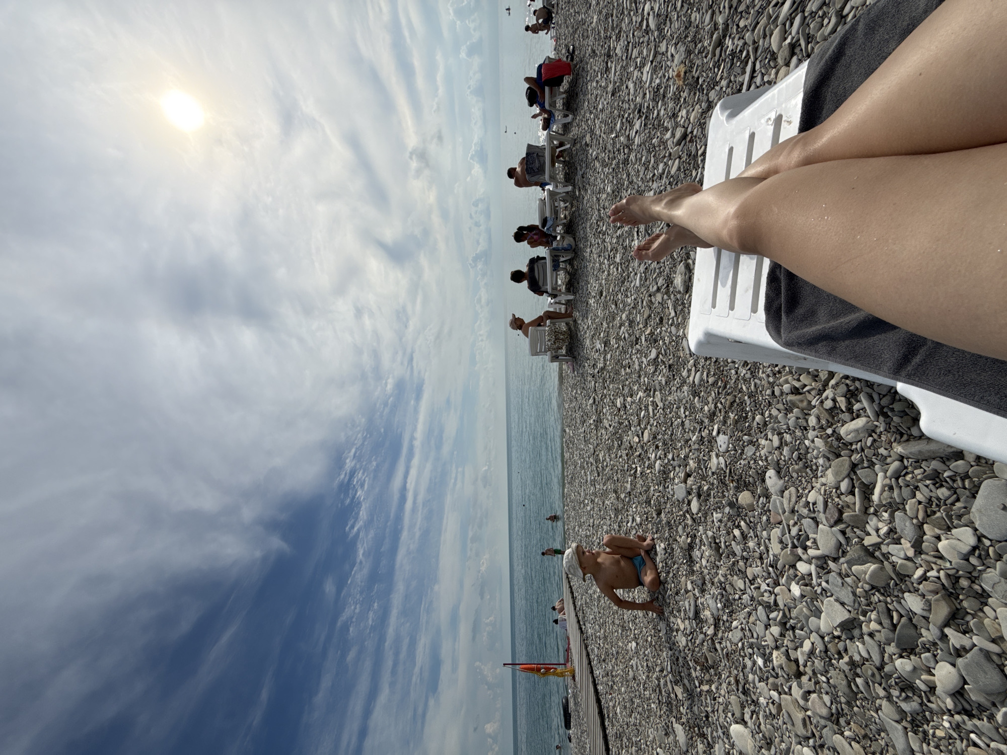 A first-person perspective shot taken from a lounge chair on a pebble beach in Nebug, Russia. The image shows the legs and feet of the person sitting in the lounge chair in the foreground. The beach extends towards the sea, with several people sitting on lounge chairs in the middle ground. The sea is calm and blue, with a few small boats visible in the distance. The sky is partly cloudy with the sun visible. A young boy in a straw hat is sitting on the pebble beach in the left middle ground. There is a wooden pier with a red flag to the left of the boy.