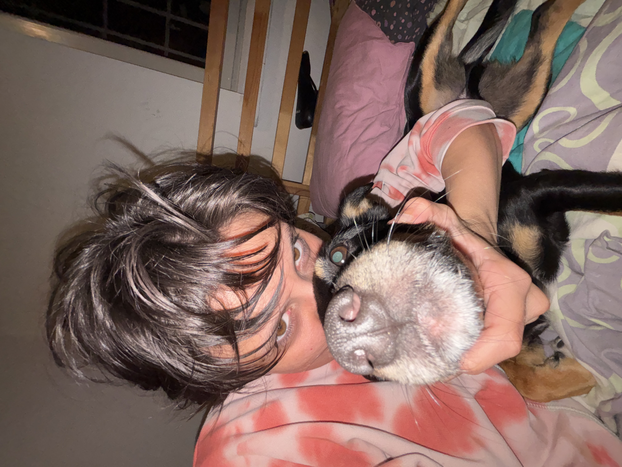 This is a close-up, indoor shot of a person and a dog, taken from a low angle. The person, with dark, unkempt hair and light brown eyes, is wearing a pink and white tie-dye sweatshirt. They are looking directly at the camera, with their face close to the dog. The dog, which appears to be a medium-sized breed with black and tan markings, is also facing the camera. Its snout is prominent in the foreground, and its eye is visible. The person's hand is holding the dog's snout. They are lying down, likely on a bed, as indicated by the presence of a pink pillow and patterned bedding in the background. The environment suggests a residential setting, possibly a bedroom. There are no visible indicators of the location being in the city of Sofia, Bulgaria.