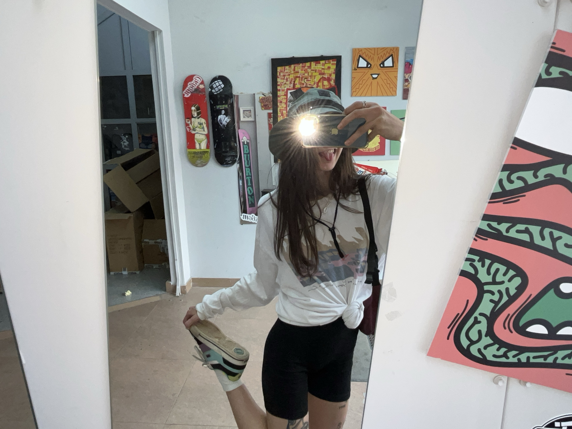 The image shows a young woman in athletic shorts and a long-sleeved shirt, taking a mirror selfie. She is wearing a hat and holding up a phone to take the picture. The background appears to be an indoor space with a wall decorated with skateboards and artwork. There are also cardboard boxes visible, suggesting a storage or work area. The environment appears to be a personal space or a creative studio. The woman is wearing sneakers and socks, and her left leg is lifted, showing off her footwear. The lighting in the image is bright, with a strong flash from the phone illuminating her face. There are no specific landmarks or details that would allow for identification of a location in Krasnoyarsk, Russia.