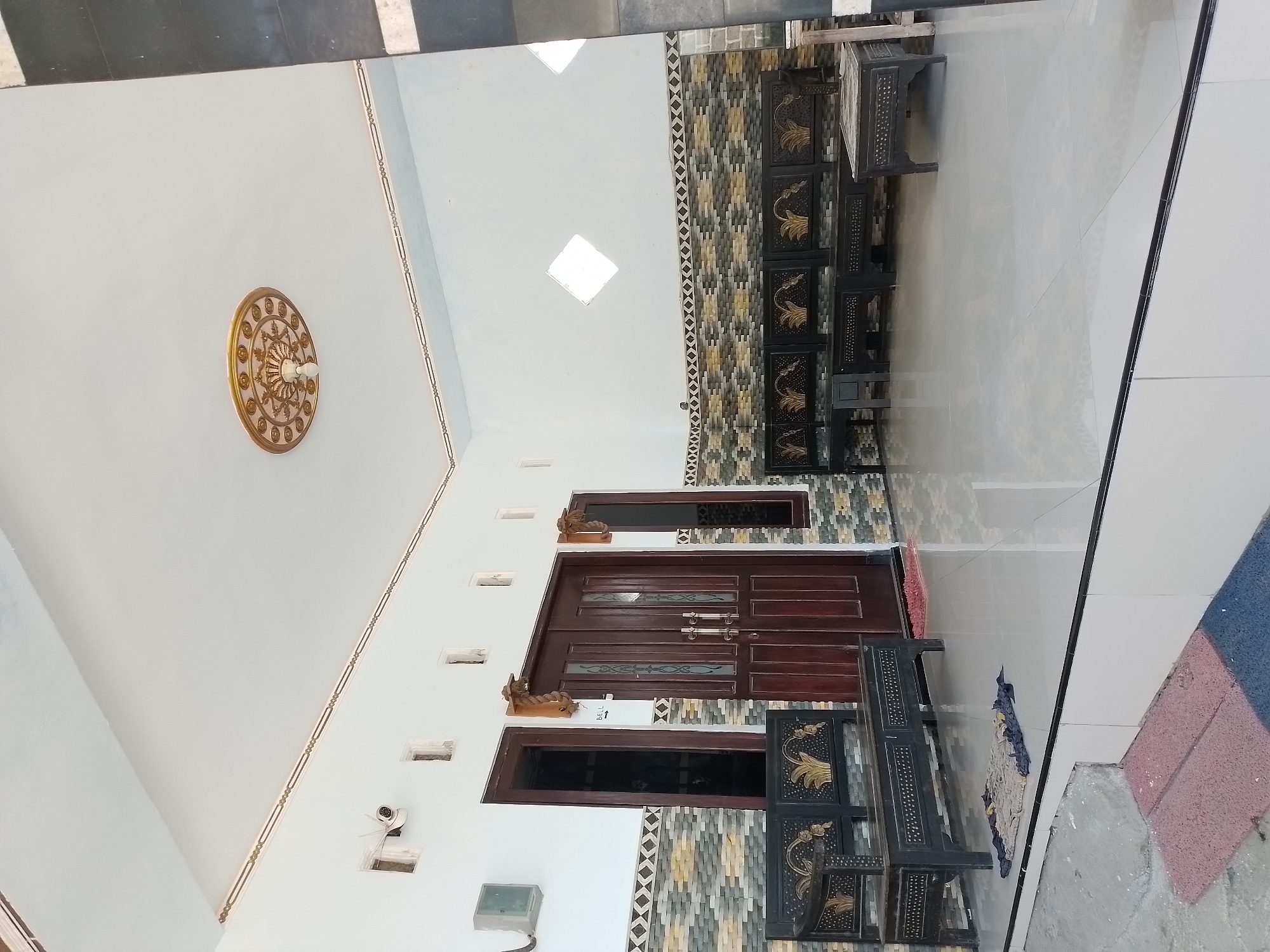 This is a photograph of the exterior of a building in Ngancar, Indonesia. The building is white with dark brown wooden double doors and a decorative gold ceiling medallion. There are black wooden benches with decorative gold detailing on the porch. The floor is tiled with a light gray color. To the left of the doors, there is a security camera mounted on the wall. Above the doors and to the right, there are small square windows. A decorative tiled pattern runs along the wall below the windows.