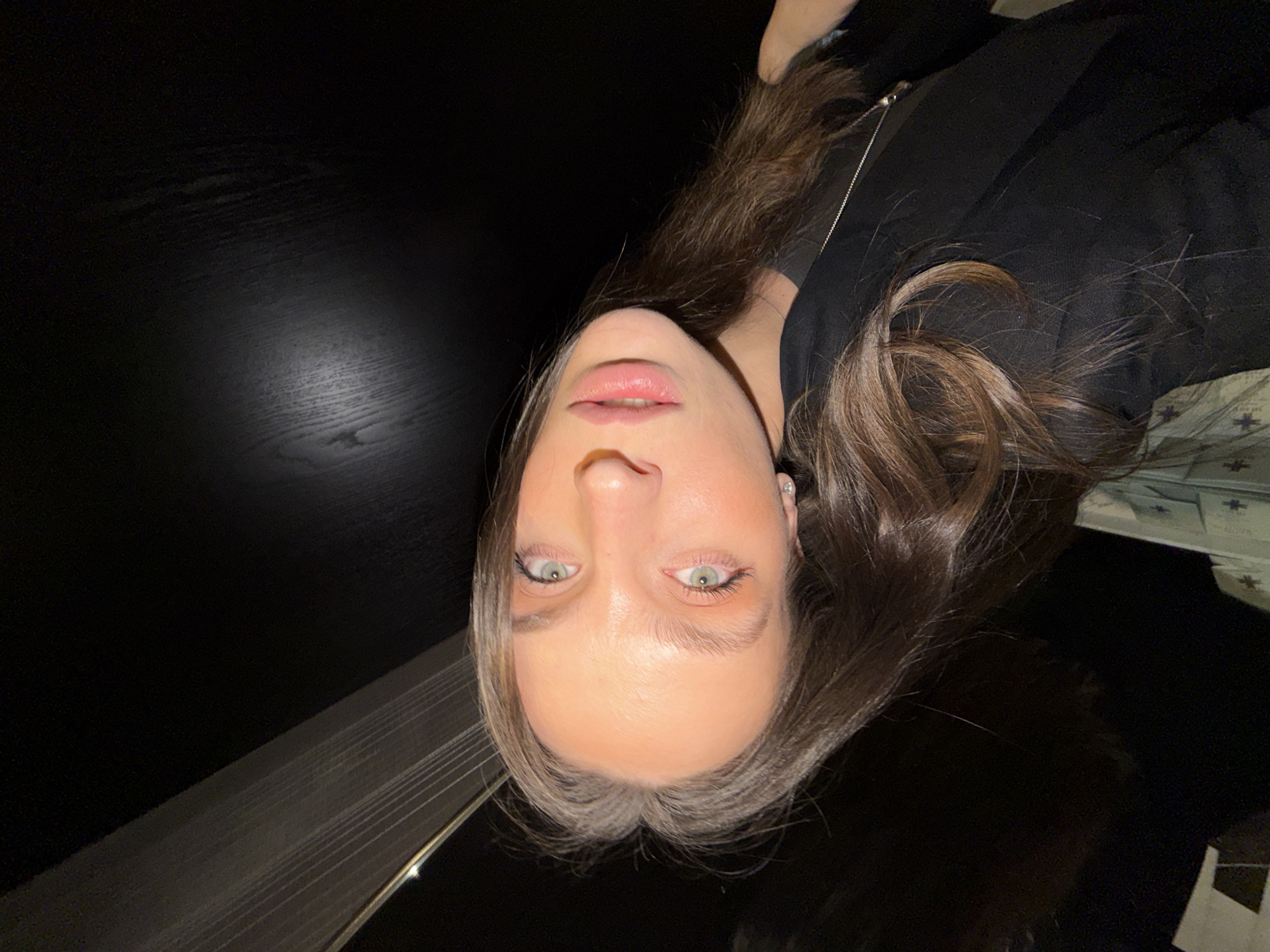 A person's face and torso are positioned in the center of the frame, tilted to the left. The person has light skin, light green eyes, and light brown hair with some gray strands visible near the hairline. The person is wearing a black top and a silver necklace. The background is mostly dark and includes a black woodgrain surface on the left, with an unclear object appearing on the upper right. The image does not provide sufficient details to identify the specific location in Budva, Montenegro.