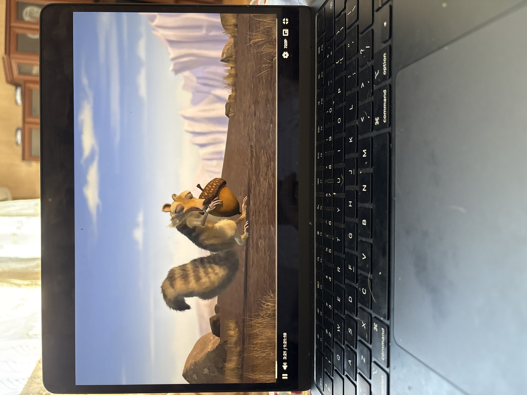 A laptop is positioned on a surface with a keyboard and trackpad visible. The laptop screen displays a scene from the animated movie "Ice Age," featuring the character Scrat holding an acorn. The background of the movie scene depicts a barren landscape with rocky formations and a pale blue sky with wispy clouds. The lower portion of the laptop screen shows playback controls, indicating the video is at 3:21 of 1:21:10 with a resolution of 720p. The overall environment suggests an indoor setting, possibly a home, with furniture visible in the background through a sheer curtain. There is no discernible location in Anton, Bulgaria associated with this image.