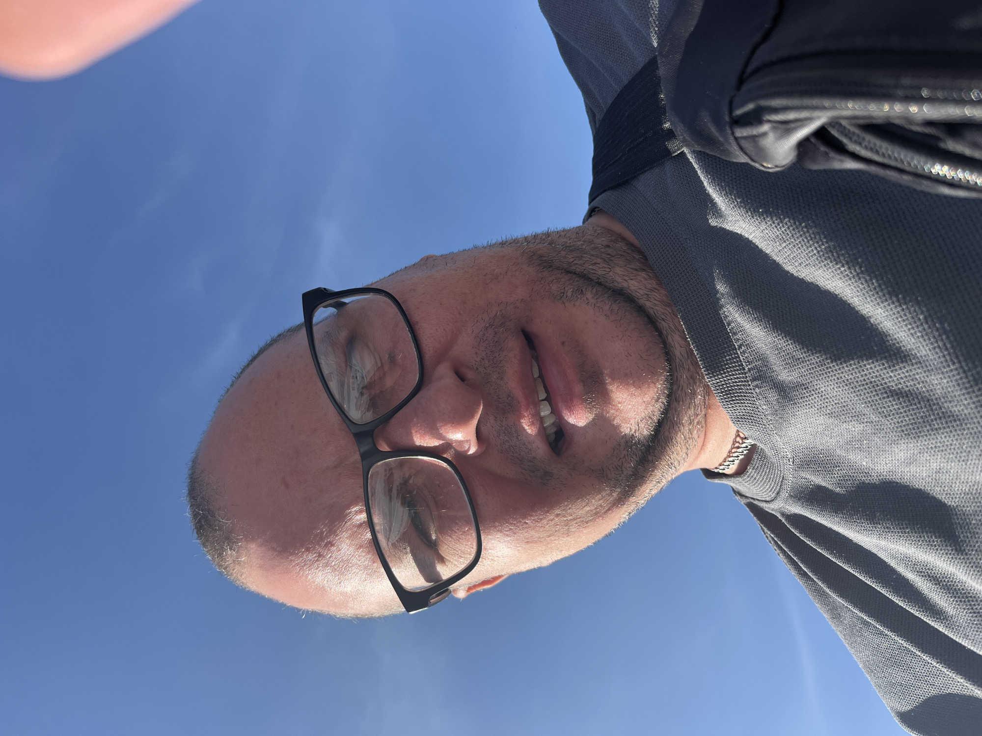 A man wearing glasses and a grey t-shirt is captured from the chest up, against a backdrop of a clear blue sky. The man's head is tilted, and he is looking towards the camera. His right hand, partially visible at the top right of the frame, appears to be holding the camera. The man has a fair complexion, a receding hairline with some grey hairs, and a light stubble. He is wearing black-rimmed glasses. He has a silver chain necklace. The man is wearing a dark-colored backpack strap over his right shoulder. The image does not provide any identifiable landmarks or clues to determine its location in Minsk, Belarus.
