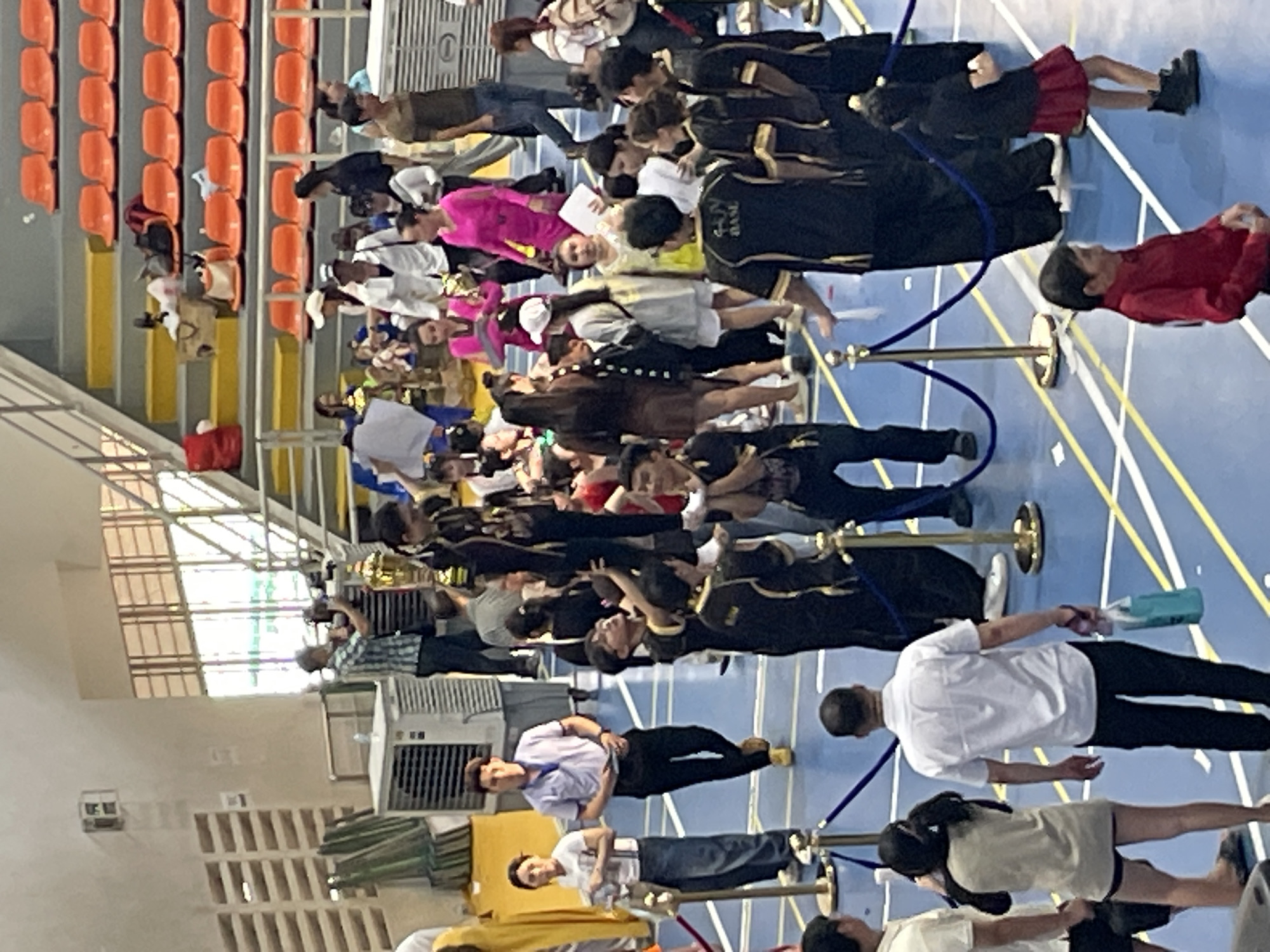 The image depicts an indoor event, likely a competition or ceremony, taking place in a gymnasium with blue flooring marked with yellow lines. In the background, orange bleachers are visible, suggesting spectator seating. A variety of people are present, many of whom appear to be participants or organizers, dressed in a mix of casual and uniform attire.
In the foreground, a man in a white collared shirt and black pants walks away from the camera, carrying a blue bag in his right hand. To his left, a woman in a beige top and dark shorts stands, her head turned away from the camera. A group of individuals dressed in black and gold uniforms are clustered together, with one person standing on the shoulders of another, creating a human pyramid. Some of these individuals are holding trophies.
Other individuals in the crowd are wearing brightly colored clothing, including a woman in a vibrant pink dress and another in a yellow dress. A man in a blue and white plaid shirt is operating a camera. The scene is cordoned off with blue ropes and gold stanchions. The presence of cooling units and the overall setup suggest an organized event within a public venue. The location is identified as Thuận An, Vietnam, implying this gymnasium is situated within the city.