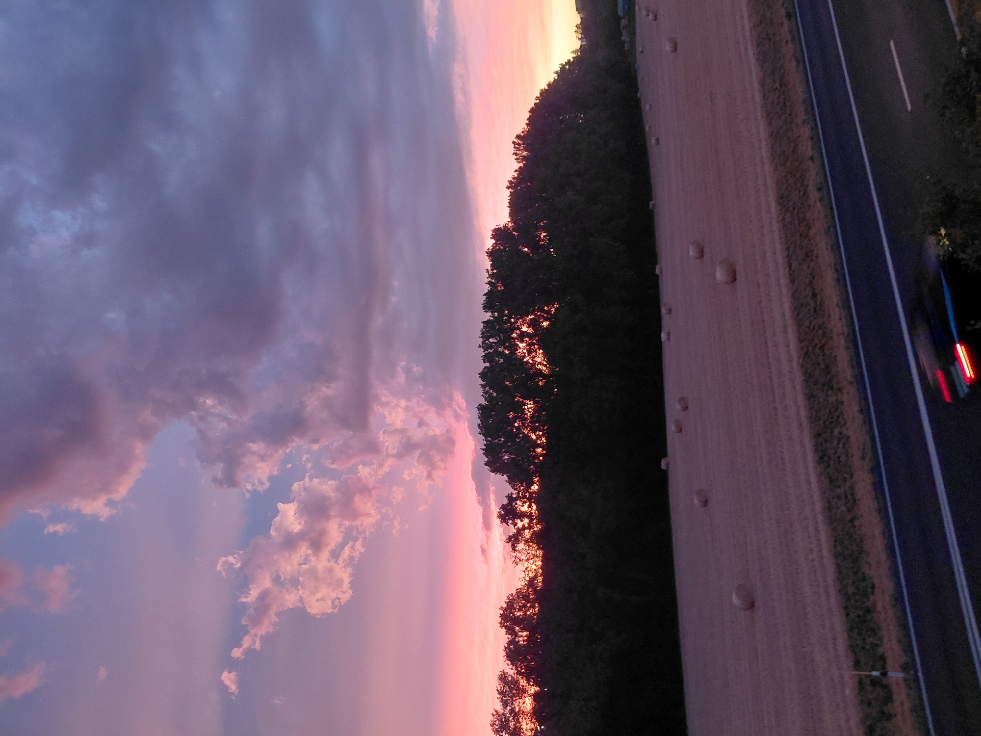 A rural landscape is depicted during sunset. The sky is a vibrant mix of pink, purple, and gray clouds. In the foreground, a field with several hay bales is visible. A line of trees runs across the middle ground, separating the field from the sky. A highway is in the lower right corner of the image, with a vehicle's taillights blurred due to motion. The scene appears to be in the vicinity of Ecublens, Switzerland, judging by the hay bales in the fields and the typical rural scenery.