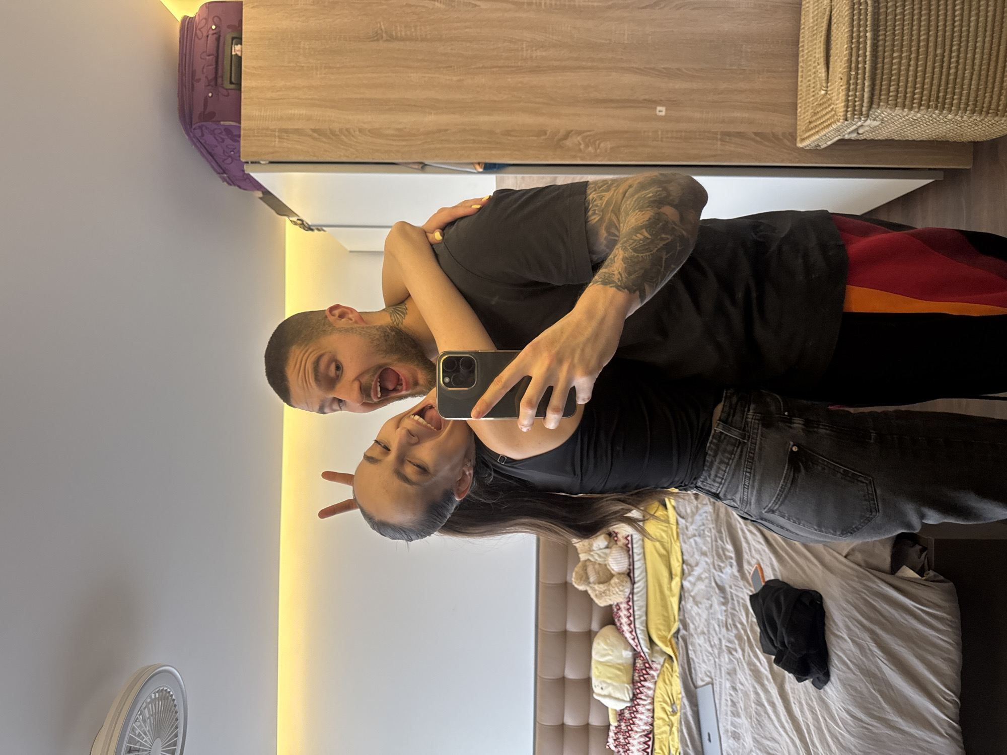 Two people are taking a selfie in a room. A man with a shaved head, wearing a black t-shirt and with arm tattoos, is holding a phone, and a woman in a black top and grey jeans is making a peace sign with her fingers. The room contains a bed with various colored blankets and pillows, a wall fan, a wardrobe, and a suitcase on top of the wardrobe. The lighting is from a light strip on the wall. There is no indication of the location of Sofia, Bulgaria.