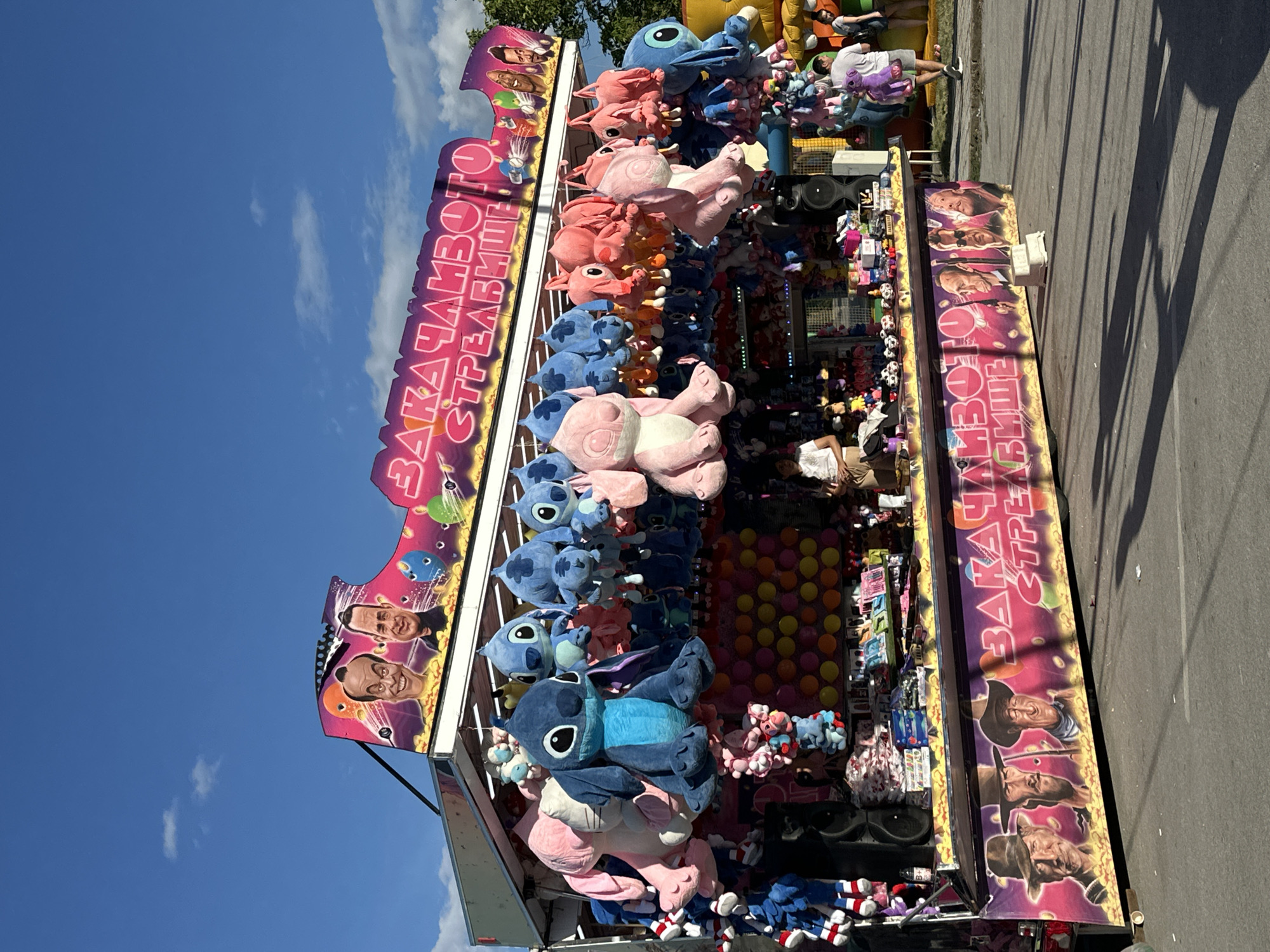 The image captures a carnival game stall in the city of Gorna Oryahovitsa, Bulgaria. The stall is adorned with colorful plush toys, primarily blue and pink Stitch characters from Disney's Lilo & Stitch. Above the toys, a banner displays caricatures of several individuals, including what appear to be political figures, and text in Bulgarian reading "ЗАКАЧАНБОТО СТРЕЛИЩЕ," which translates to "The Fun Shooting Range." Another banner at the bottom of the stall features painted portraits of three cowboys, reminiscent of classic Western film stars. In the background, people are visible, some walking by and others manning the stall. The sky is blue with scattered clouds, and the overall setting suggests a bright, sunny day at an outdoor amusement event.