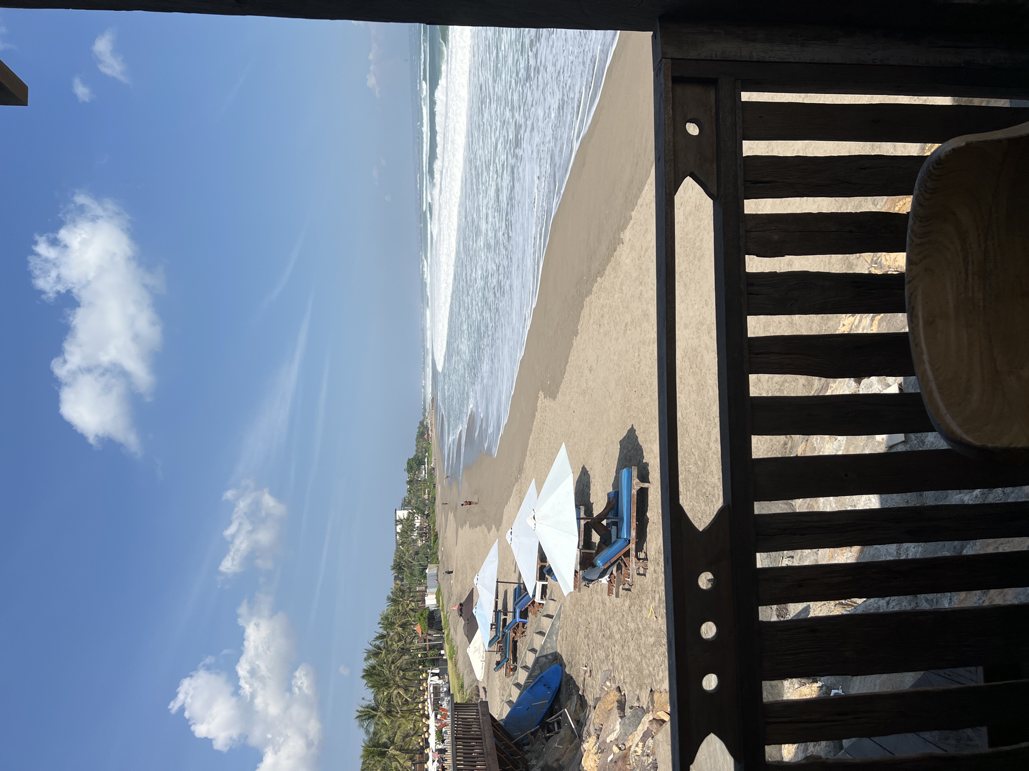 An outdoor, eye-level, medium shot captures a view of Seminyak Beach in North Kuta, Indonesia, looking out towards the ocean. The foreground features a wooden railing with vertical slats and decorative cutouts, framing the scene. Beyond the railing, a sandy beach is dotted with several blue lounge chairs and white and blue umbrellas, some occupied by individuals. To the left, a wooden deck or building is visible with more seating and umbrellas, and a blue surf or paddleboard leans against a rocky embankment. Palm trees line the beach in the distance, interspersed with buildings. The ocean is active with white-capped waves rolling onto the shore under a bright blue sky with scattered white clouds.