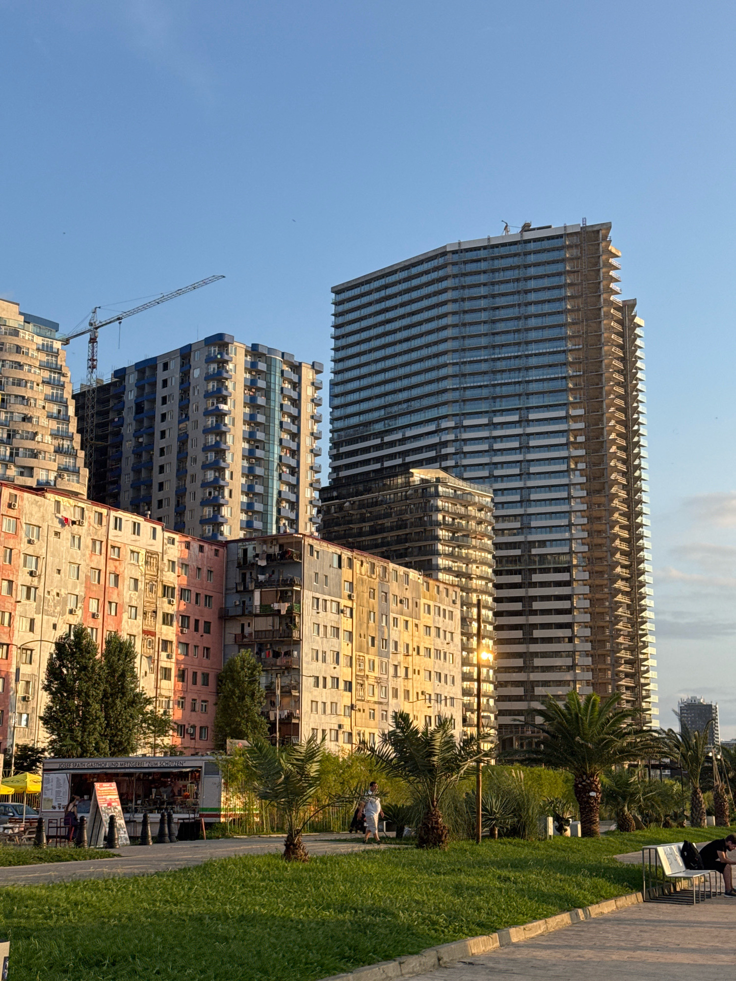 The image depicts a waterfront promenade in Batumi, Georgia, with a clear blue sky overhead and the golden hour sun casting long shadows. In the background, a cluster of buildings of varying architectural styles and construction stages dominate the skyline. A prominent, unfinished high-rise with a glass facade and exposed concrete balconies stands tall on the right. To its left, a blue and gray apartment building with numerous balconies is situated, with a construction crane visible above it. Further left, older, multi-story buildings painted in pastel colors like pink and beige, with visible air conditioning units and laundry hanging out, line the street. A food truck with text in German, "JOSEF SPATH GASTHOF UND METZGEREI ZUM SCHÜTZEN," is parked near the older buildings. The foreground features a lush green lawn, with several palm trees and other vegetation. A paved walkway runs along the waterfront, where a few individuals are seen strolling and sitting on a bench. The overall scene captures the juxtaposition of new development and older urban structures in a coastal city.