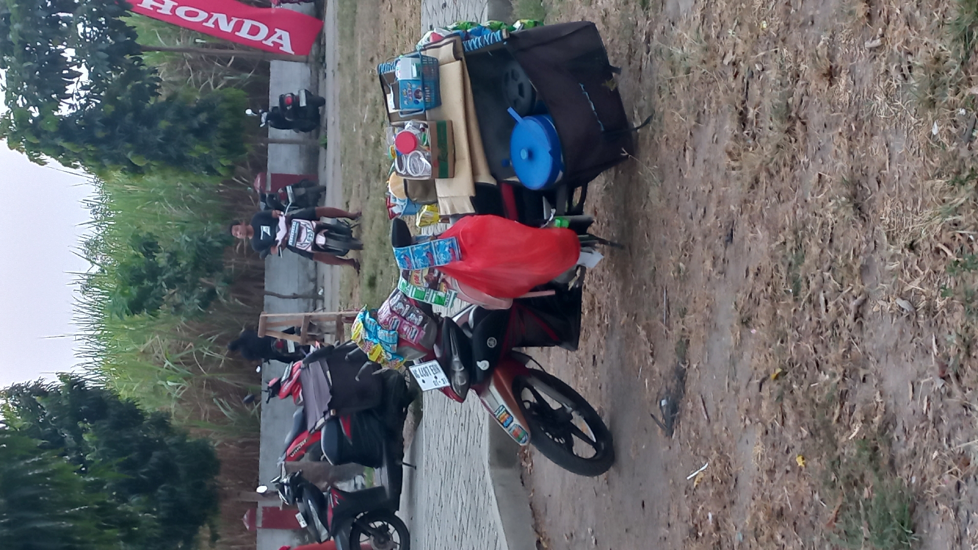 A motorcycle is parked on a dirt and grassy area. The motorcycle is loaded with various items, including bags of snacks, a red plastic bag, a blue plastic container, and other packaged goods. A man is seated on another motorcycle in the background. A red and white Honda banner is visible to the right. The environment appears to be an outdoor area with trees and tall grass in the background. The scene is likely located in the city of Pare, Indonesia, given the presence of a Honda banner, a common brand in the region.
