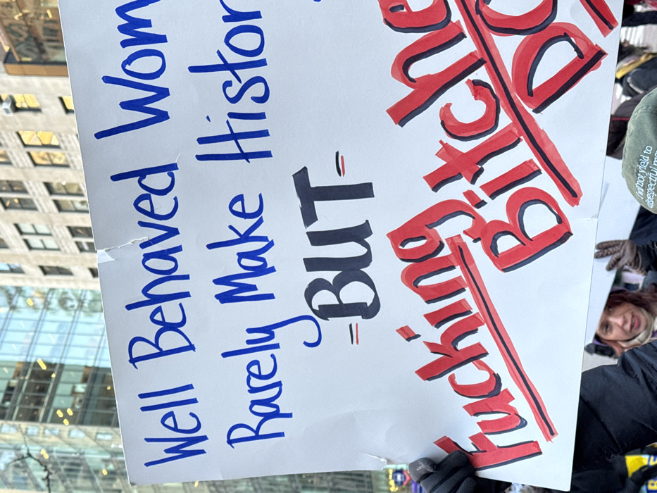 A protest sign reads "Well Behaved Women Rarely Make History - BUT - Fucking Bitch". The last two words are in red and struck through with a line. The words "Well Behaved Women Rarely Make History" are in blue. The sign is being held by someone outside in front of buildings. A person wearing a black coat and gloves is visible in the foreground. Another person is in the background wearing an olive drab jacket and another person with red hair is visible behind.