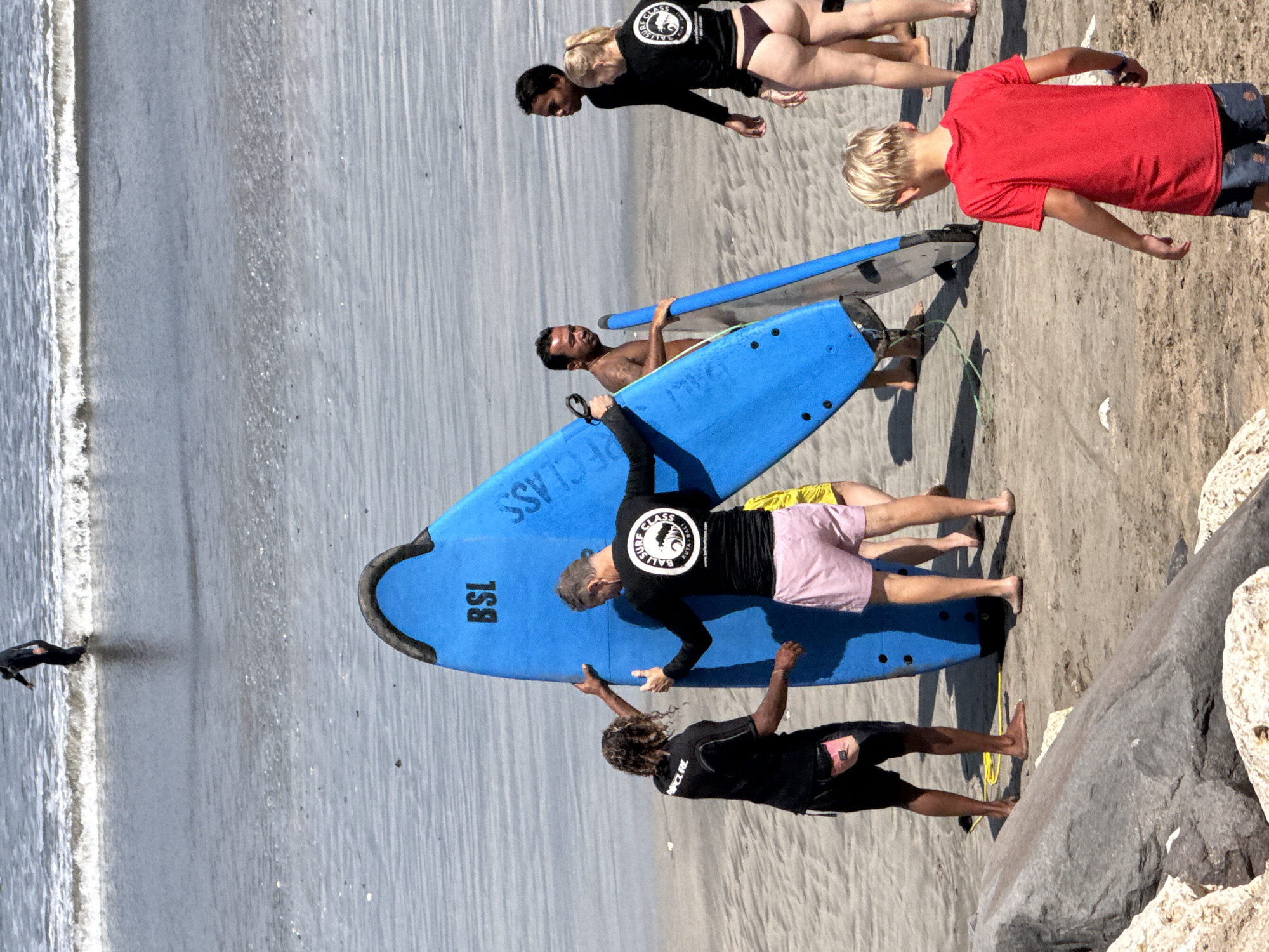 The image depicts a group of people on a sandy beach in Kuta, Indonesia, with surfboards. A young boy in a red shirt and dark patterned shorts is standing, looking down. Beside him, another person wearing a black rash guard with a circular logo and pink shorts is lying on a blue surfboard. Another person in a black rash guard is kneeling on the same blue surfboard. Further down the beach, a man with dark hair is lying on a surfboard. In the background, two people in black tops and dark bottoms are standing on the sand, and another person is partially visible lying on a surfboard. The sea is visible on the left side of the image, with waves breaking on the shore. There is visible text on the blue surfboard: "BSL" and "CLASS". The weather appears sunny, suggesting it is daytime. The overall scene suggests a surf lesson or a group outing to the beach.