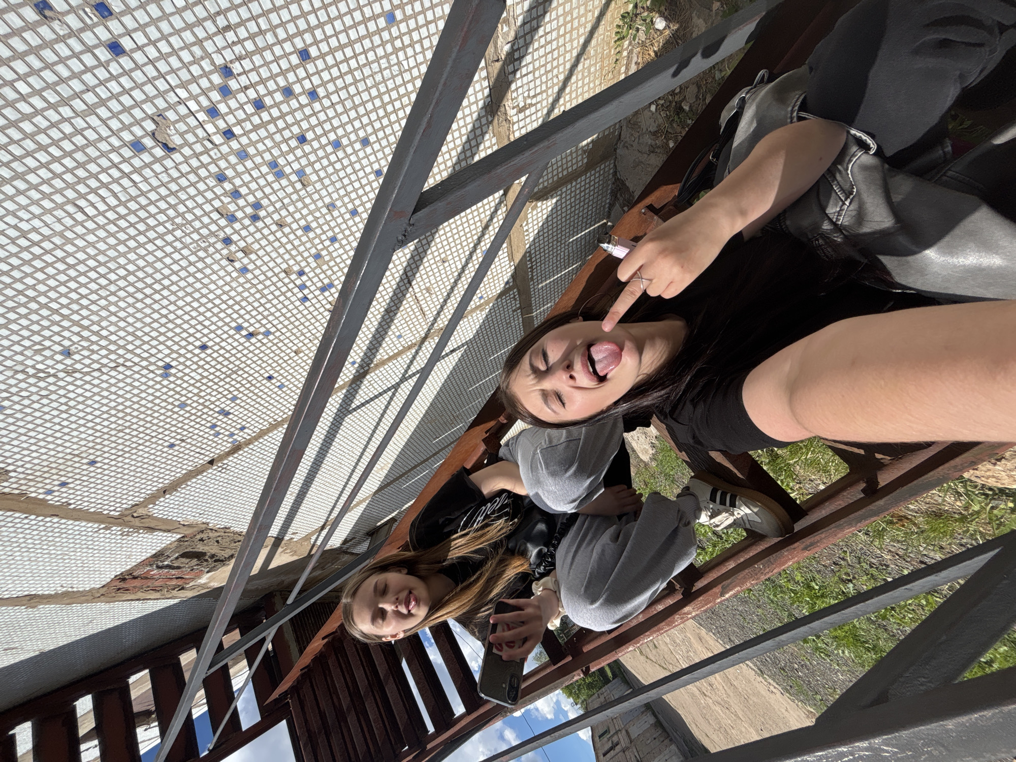 Two young women are sitting on a metal staircase, possibly in Chaikovsky, Russia. The staircase has a mesh ceiling above. One woman holds a smartphone, while the other sticks out her tongue and points to it. They are both wearing casual clothing. The background includes greenery and a building exterior.